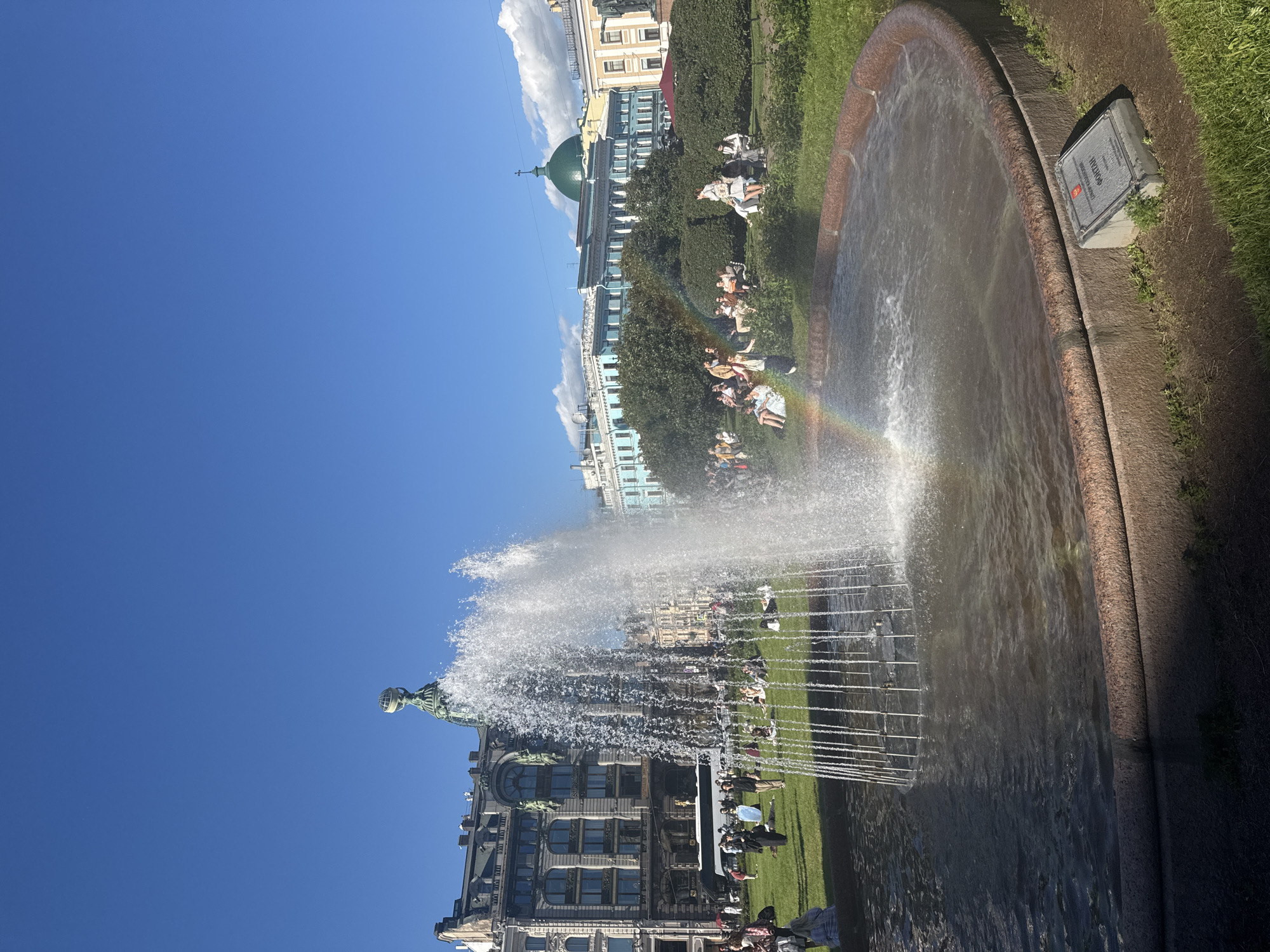 This image depicts the Singer House (Dom Knigi) and the nearby fountain in Saint Petersburg, Russia, on a sunny day. A large, multi-tiered fountain is in the foreground, with water jets creating a mist. A faint rainbow is visible within the spray of the fountain. In the background, the Singer House, an iconic Art Nouveau building, stands tall with its distinctive glass dome topped by an eagle statue. Several people are scattered across the green lawn surrounding the fountain, some sitting, some standing, enjoying the pleasant weather. Lush green trees and other historic buildings with distinct architectural styles, including one with a green dome and a cross, line the horizon. The sky is a clear blue with a few wispy clouds. A plaque is visible in the lower right corner of the image, in front of the fountain's basin.