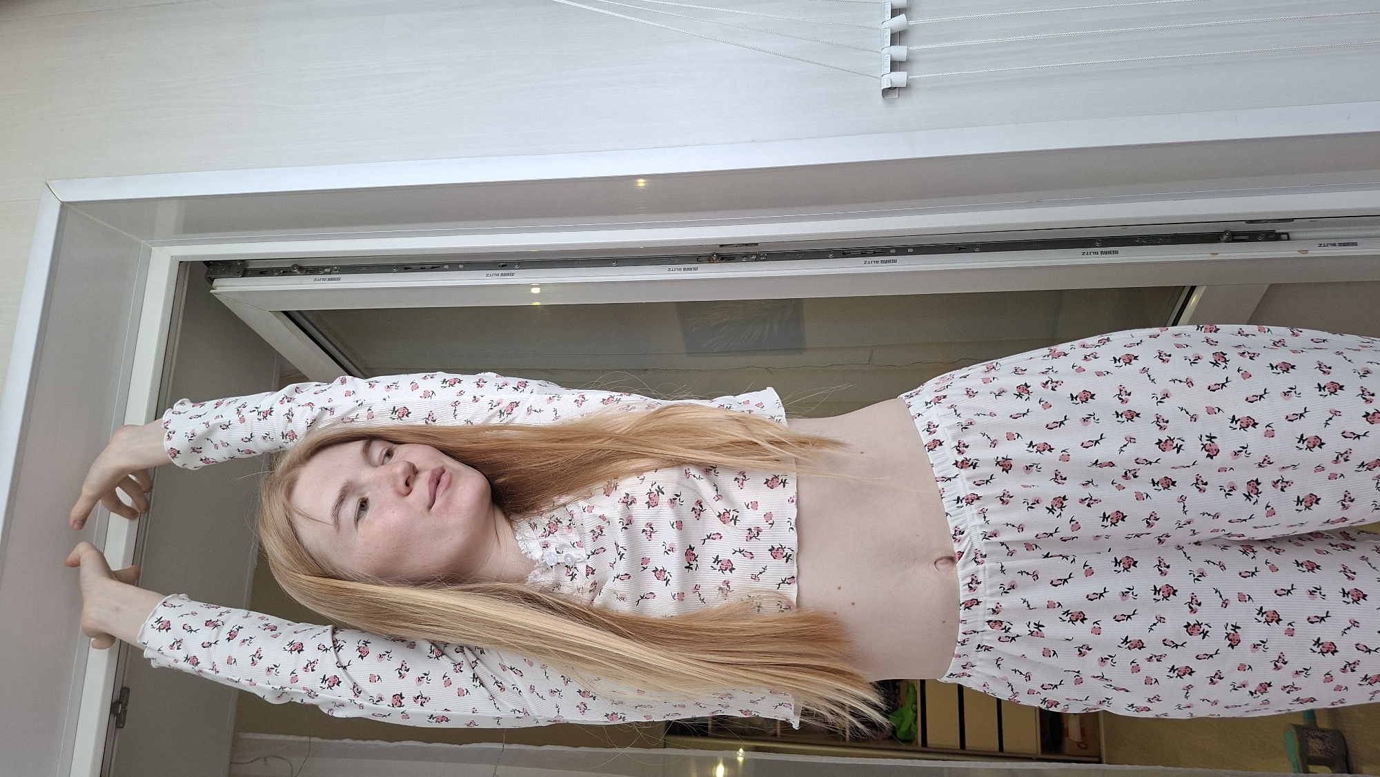 A woman with long, straight, light brown hair is lying down, positioned vertically in the frame, against a backdrop that appears to be an open window or door. She is wearing a matching two-piece loungewear set, featuring a white fabric with a repeating pattern of small pink and black flowers and green leaves. The set consists of a long-sleeved crop top and long pants. Her arms are raised and extended outwards, with her hands resting on the white frame of the opening. Her head is tilted slightly back, and she is looking upwards towards the viewer with a relaxed expression. Her belly button is visible.
Behind her, through the open frame, there are reflections on what looks like a glass pane. Above her, what appears to be a white window blind with horizontal slats and cords is visible. The immediate surroundings are light-colored walls or frames, suggesting an indoor setting, possibly a room in a house in Chaikovsky, Russia. The light in the image suggests it is daytime, although the exact time of day or weather cannot be determined.
Visible text includes "REHAU BLITZ" repeated multiple times on what appears to be the window mechanism or frame.