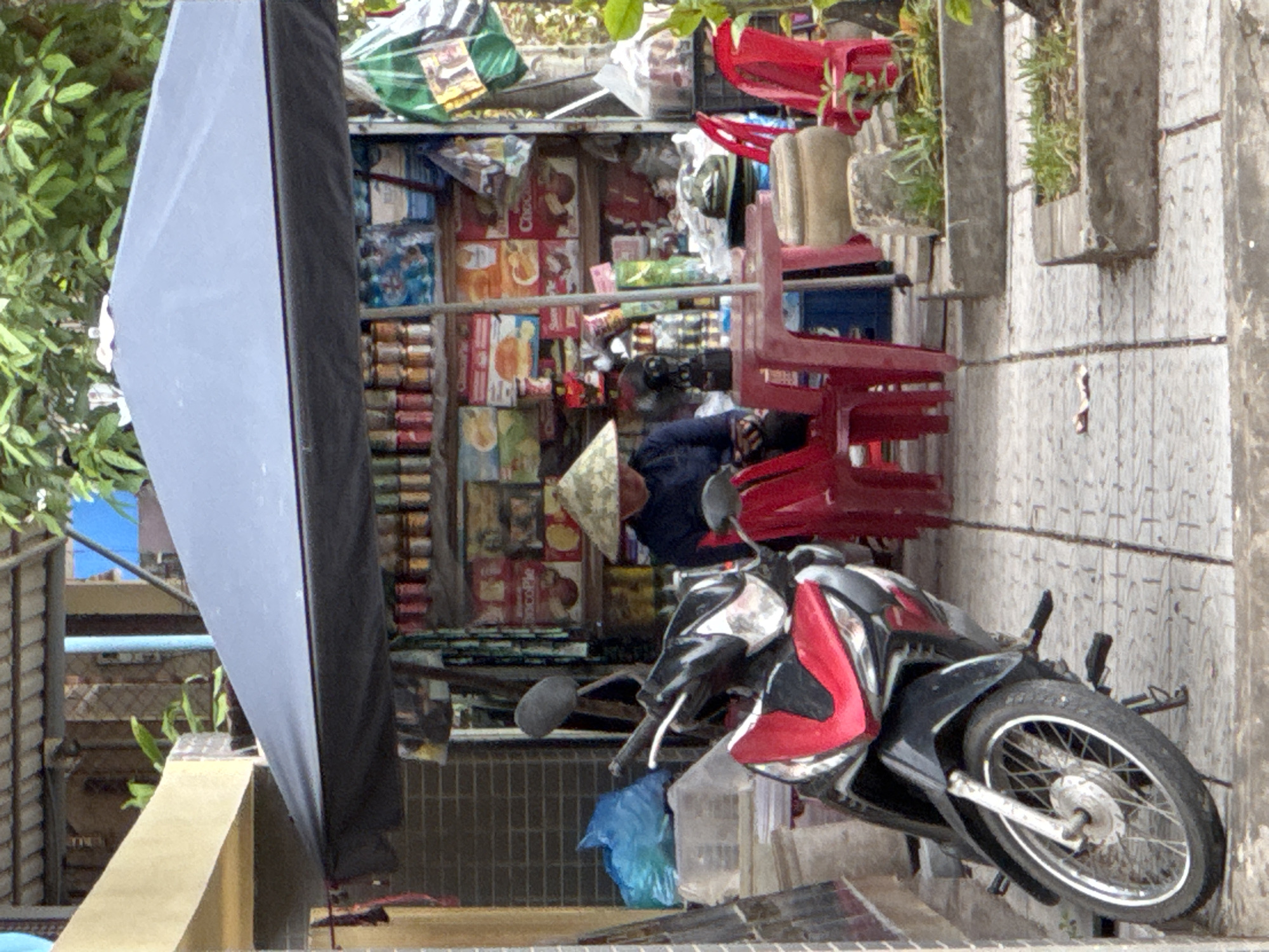 A street stall in Đông Hà Ward, Vietnam, is set up under a dark blue umbrella. A person wearing a conical hat is seated behind the stall, which is filled with various packaged goods, including cans, boxes, and bags. In front of the stall, a red plastic table and three red plastic chairs are arranged. To the left of the stall, a black and red motorcycle is parked. The ground is paved with square tiles. Greenery is visible in the background, including trees and plants.