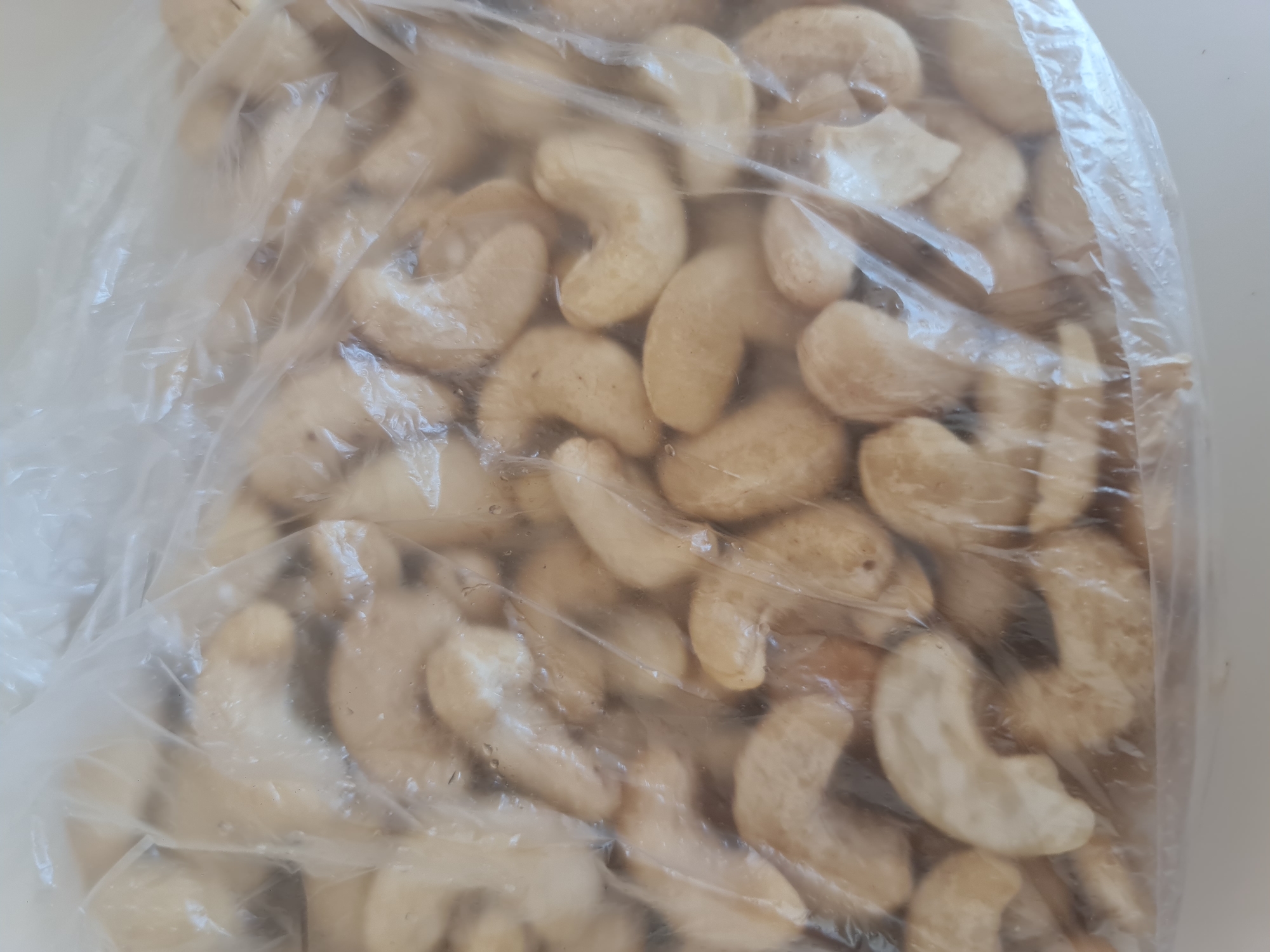 The image shows a close-up, overhead view of a clear plastic bag filled with whole, raw cashews. The cashews are a light tan color and have the characteristic curved shape. Some of the cashews are slightly uneven in color, with darker spots or markings. The plastic bag is slightly wrinkled and shows some condensation on its inner surface, indicating the cashews might be fresh or have been stored in a humid environment. The lighting is bright and diffused, making it difficult to discern any specific environmental details. There are no people or actions depicted in the image. The image is not specific to Angarsk, Russia; it is a generic depiction of a bag of cashews.