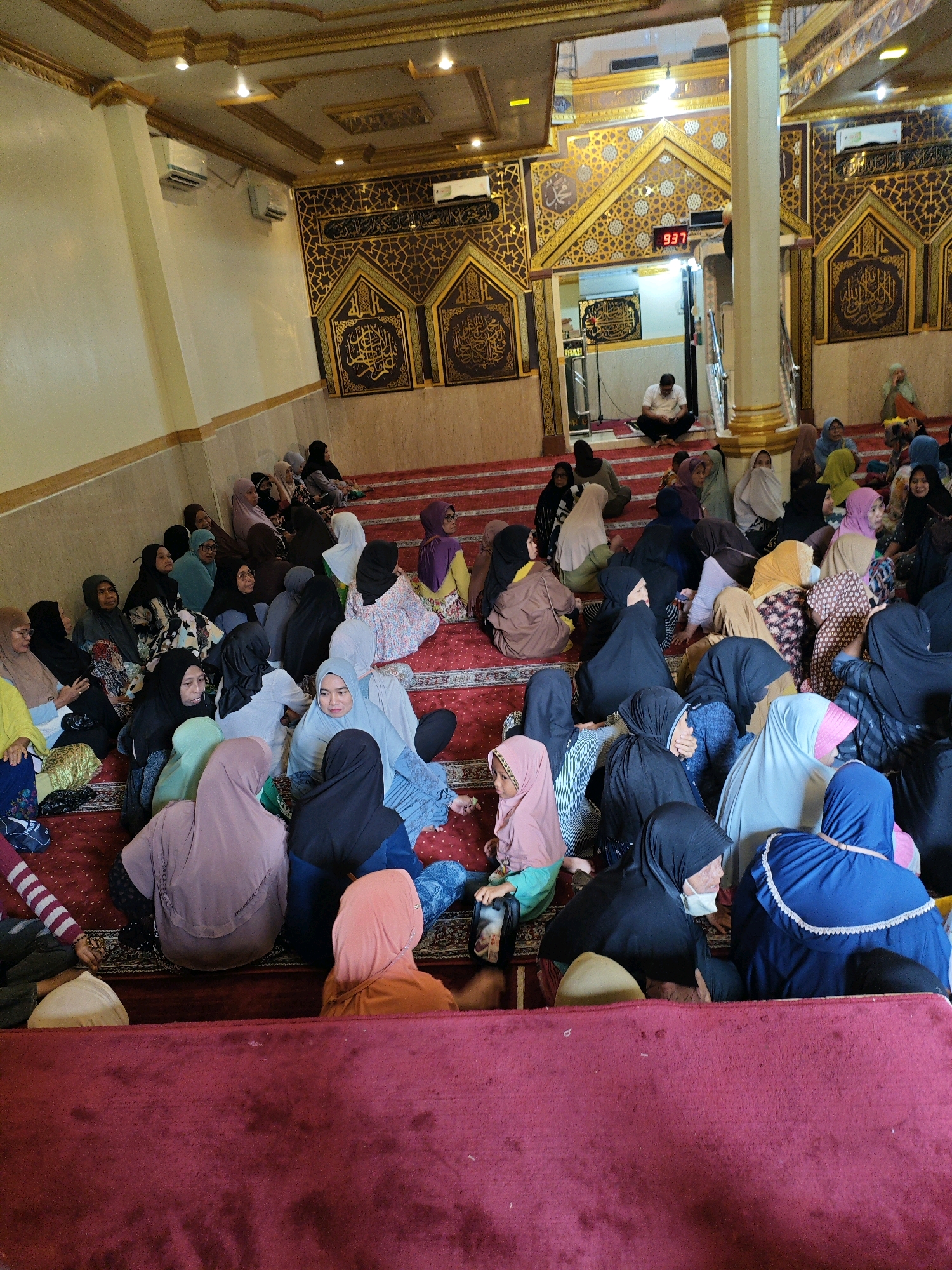The image depicts a gathering of people, predominantly women wearing headscarves, seated on a red carpeted floor inside a room with ornate Islamic decor. The room appears to be a mosque or a similar religious or community space, indicated by the patterned walls, golden accents, and calligraphy. A digital clock displaying "937" is visible on a wall. Several individuals are facing forward, suggesting they are listening to a speaker or observing an event. One man is seated in the background near an entrance. The overall scene is one of communal gathering and possibly a religious service or lecture.