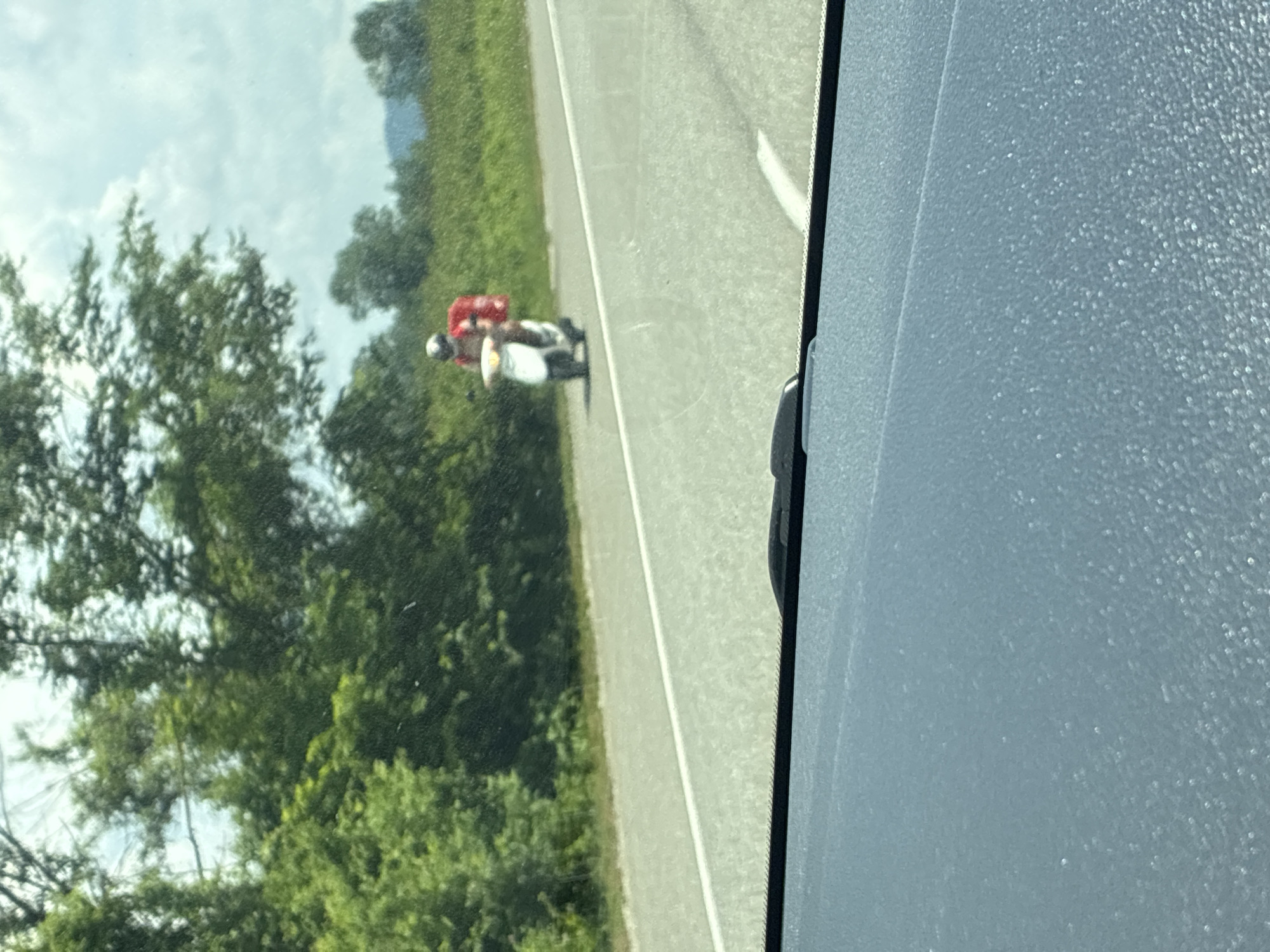 An out-of-focus image taken from inside a vehicle shows a person on a scooter driving on a paved road. The person is wearing a red shirt and a helmet, and has a red backpack on the back of the scooter. The road is bordered by green grass and trees on the left side. The sky is cloudy. The location is estimated to be in Tekos, Russia, based on the provided context, although the visual elements themselves do not confirm this specific location. The foreground of the image shows a portion of the vehicle's dashboard.