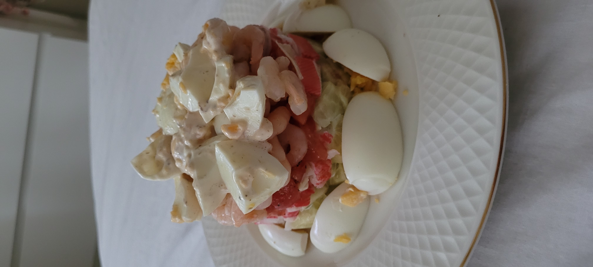 A stacked seafood salad is presented on a white plate with a gold rim, set against a white tablecloth. The salad consists of layers of chopped ingredients, including what appears to be avocado, tomatoes, and imitation crab meat, topped with shrimp and pieces of hard-boiled egg coated in a creamy dressing. Several halves and quarters of hard-boiled eggs are arranged around the base of the salad on the plate, with a scattering of crumbled egg yolk. The background is blurred, showing a white surface and a hint of a light-colored object. There is no indication of the city of Riga, Latvia, or any specific location within it.