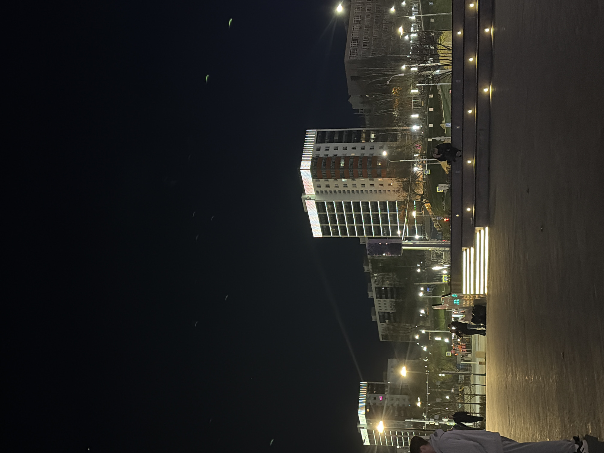 At night, the image depicts a scene in Perm, Russia. Several tall buildings are lit up, with visible windows. Streetlights illuminate the area. In the dark sky, there are several green, oval shapes, resembling balloons. People are seen walking on the sidewalk. The foreground is a concrete surface, possibly a promenade.