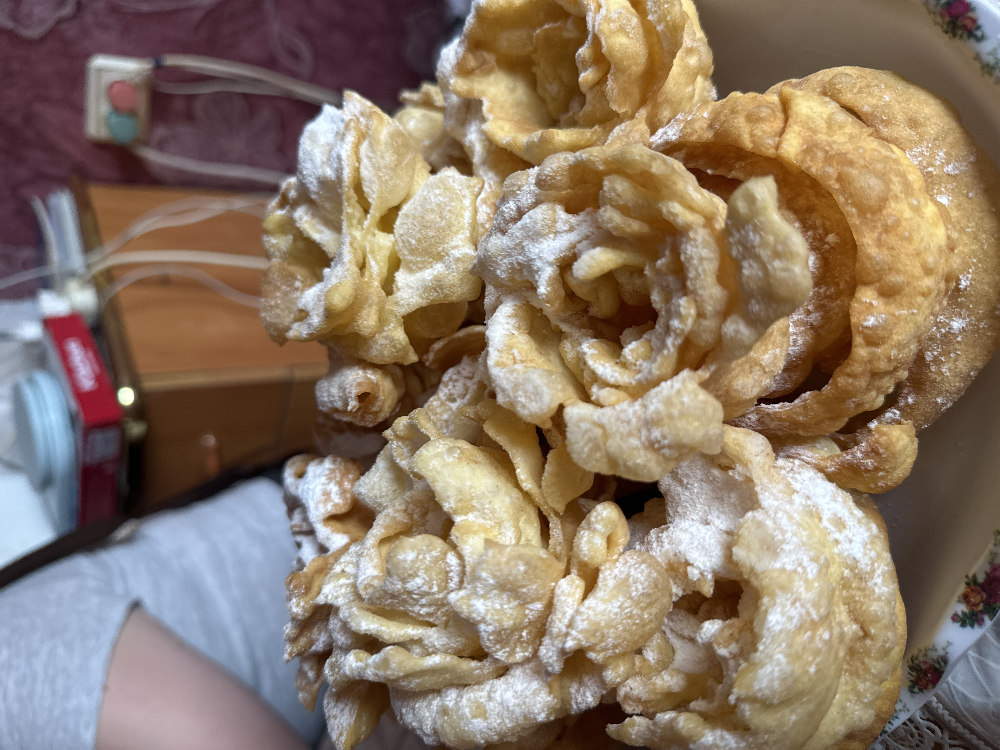 The image shows a close-up of a pile of angel wings cookies dusted with powdered sugar. The cookies appear light golden in color and are arranged in a bowl with a floral pattern. The background is slightly blurred, showing parts of a light gray garment, a wooden surface, and a wall with a light switch and outlet. The location, Коноваловка, Russia, cannot be independently verified from the image content.