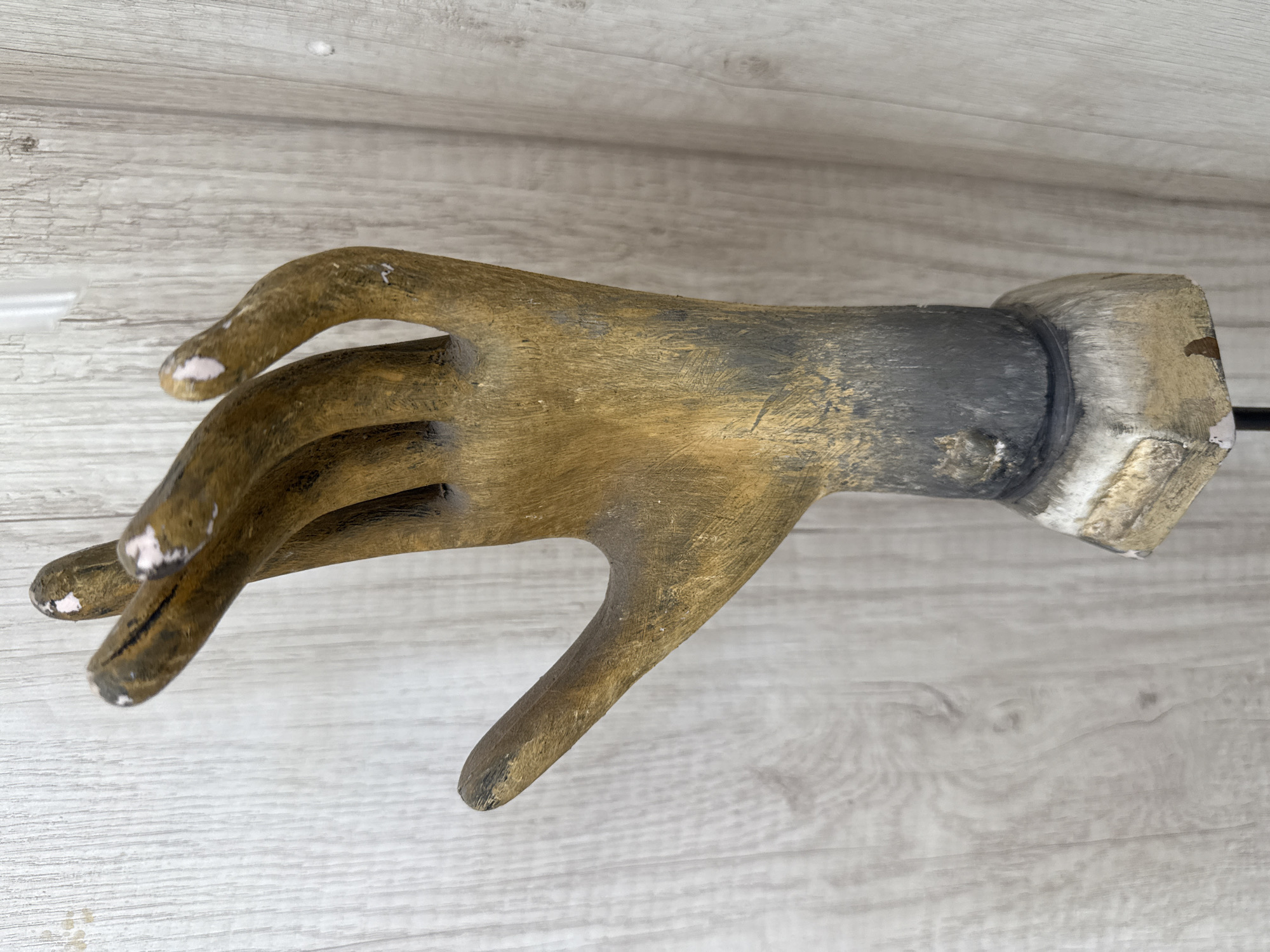 An overhead shot displays a weathered hand sculpture resting on a light-colored, wood-grain surface. The sculpture, likely made of wood or a similar material, is positioned horizontally across the frame. It depicts a right hand with fingers slightly curled and spread apart. The hand's primary color is a medium to golden-brown, showcasing a rustic and textured finish with visible grain.
The wrist and forearm section of the sculpture transition to a darker, grayish-black color, with some lighter, almost silver or white, accents on the cuff-like portion of the wrist. The fingertips show areas of wear or intentional distressing, revealing lighter, pale pink or white patches. Fine black lines are also visible on some of the fingers. A black rod, possibly a stand attachment, extends from the far end of the wrist.
The background consists of a simple, light greyish-brown wooden surface, characterized by horizontal wood grain patterns. The even lighting suggests an indoor setting. The image is a static display of an art object, with no discernible activity or event taking place. There are no visible emotions or text. While the location context is Puebla, Mexico, the object itself does not provide specific visual cues to this geographical location.