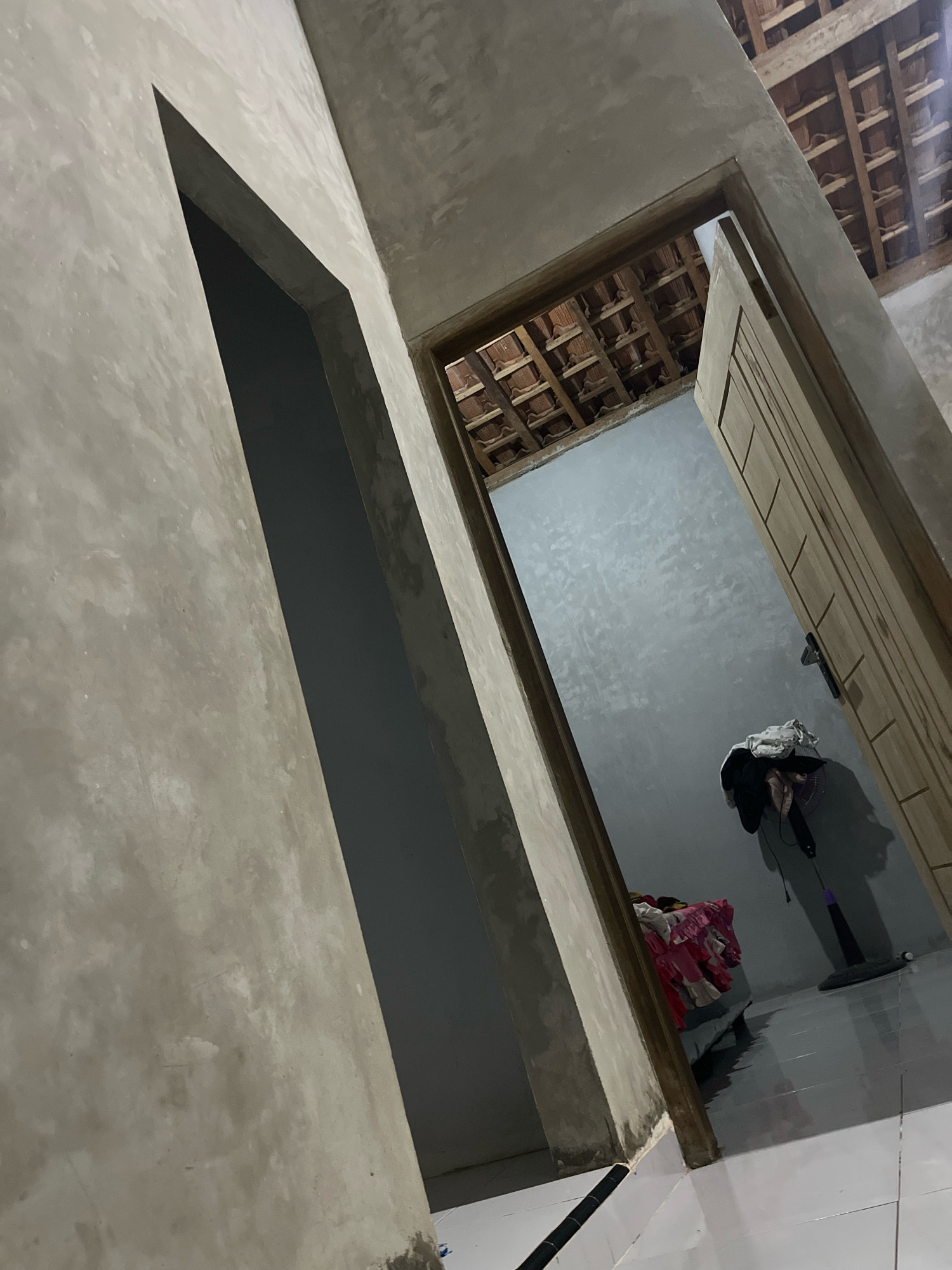This is a photograph taken in Kebumen, Indonesia, depicting an interior view of what appears to be a partially finished or under-renovation residential space. The dominant elements are unfinished concrete walls with a smooth, gray finish, and wooden door frames. One doorway, on the left, is an abstract, angled opening. Another doorway, on the right, frames a view into an adjacent room. This room has a grey concrete wall and a visible ceiling structure made of wooden beams and terracotta tiles. On the floor of this room, a standing fan is positioned near a pile of clothes. The floor in the foreground is tiled in white. The lighting suggests it is daytime, but the interior is dimly lit, casting shadows. There are no people visible in the image. The scene conveys a sense of construction or domestic activity in progress.