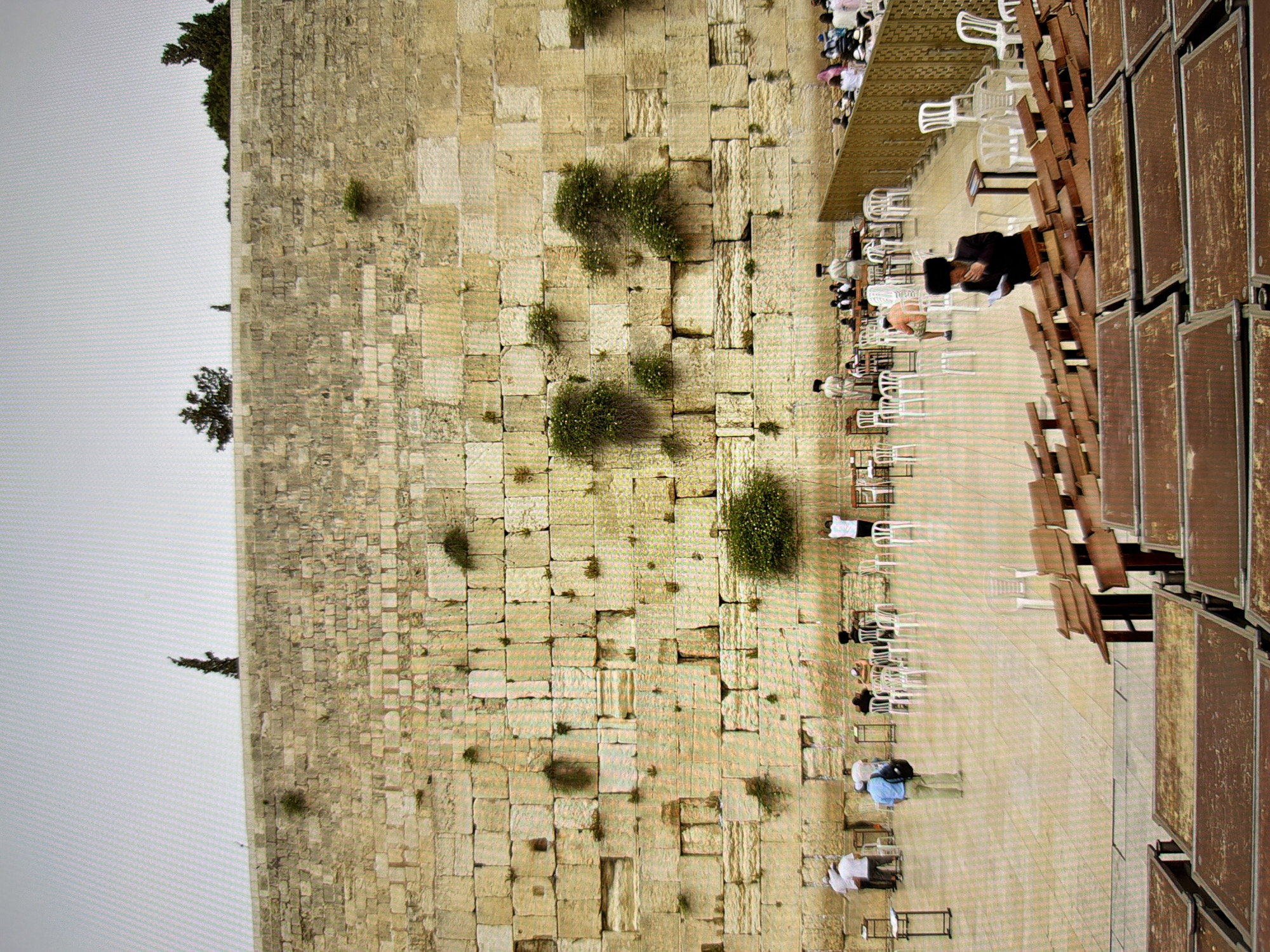 The image shows a stone wall with several people gathered in front of it. The wall is made of large, rough-hewn stones of varying shades of tan and yellow. There are plants growing out of some of the crevices in the wall. In the foreground, there are rows of wooden benches, some of which are occupied by people. In the middle ground, there is a paved area where more people are standing and sitting on chairs. Some people are dressed in traditional Jewish attire, including hats and long coats. The sky is overcast and a pale gray. This is the Western Wall in Jerusalem, Israel, not Kyiv, Ukraine.