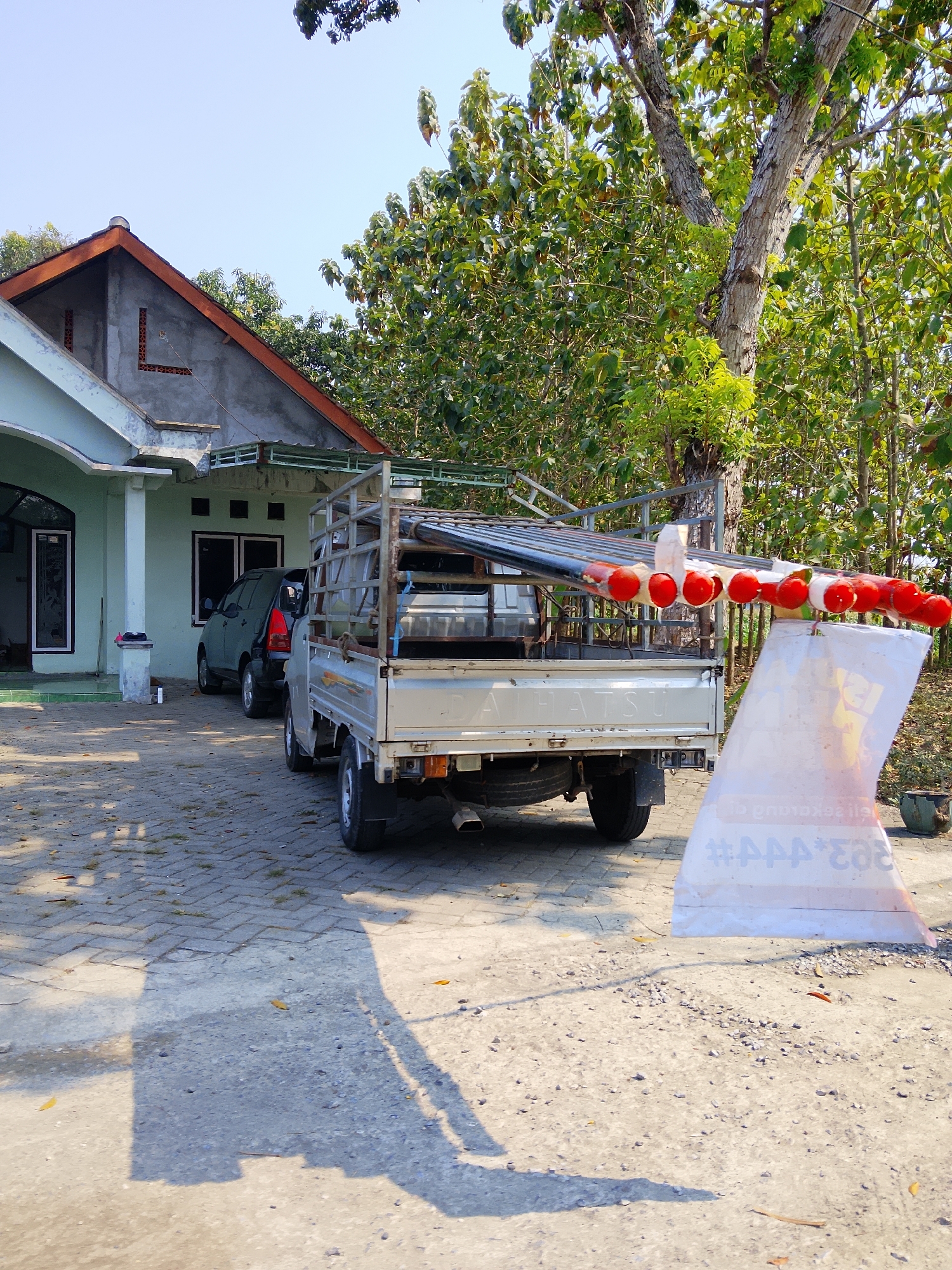 In the city of Ngaglik, Indonesia, a residential scene unfolds under a clear sky. A light-colored, single-story house with an orange-tiled roof and gray concrete walls dominates the left side of the frame. In front of the house, a dark-colored car is parked. To the right of the house, a silver pickup truck is parked. The truck is loaded with long, cylindrical objects wrapped in red and white material, possibly for construction or decoration. A large white banner with some text, partially obscured, is leaning against the truck and extends onto the ground. The ground is paved with interlocking concrete blocks in front of the house, transitioning to a dirt and gravel surface in the foreground. The scene is framed by lush green trees with dense foliage, characteristic of a tropical environment. Sunlight casts long shadows across the paving and dirt, indicating a time of day when the sun is at an angle.