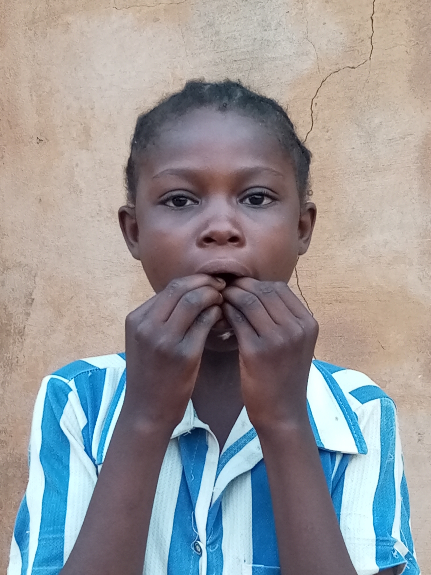 A young Black child, likely a girl given the hairstyle, is shown from the chest up, standing in front of a rough, tan-colored wall with a prominent vertical crack. The child has dark skin and braided hair. She is wearing a blue and white vertically striped collared shirt. Her hands are raised to her mouth, and she appears to be holding something between her fingers and bringing it towards her lips, as if eating or examining a small object. The lighting is natural and appears to be daylight, casting soft shadows. The background wall suggests an outdoor setting, possibly in a rural or less developed area like Tempure, Nigeria.