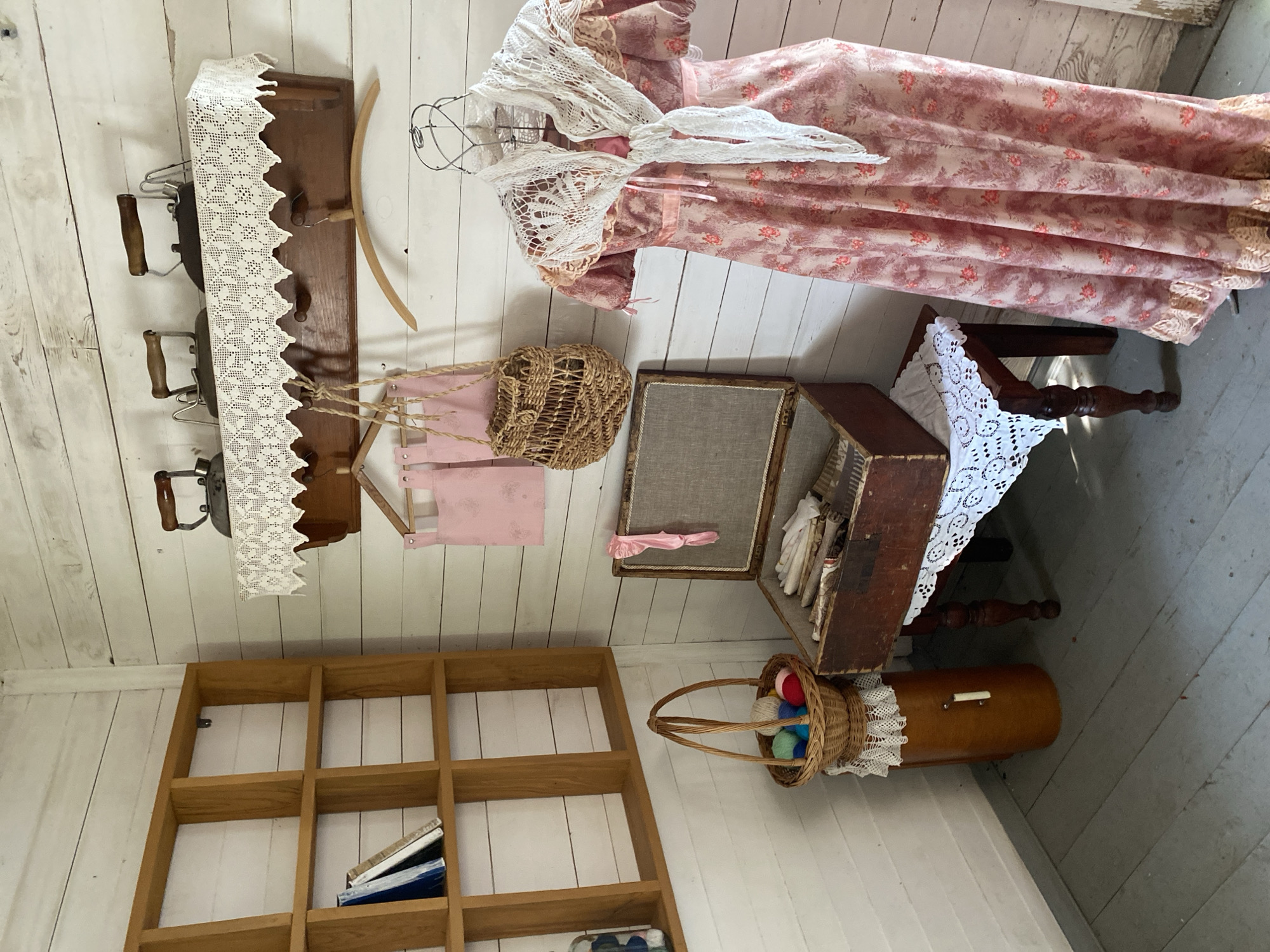 The image depicts a corner of a room, likely an exhibit in a museum, showcasing historical domestic items. The walls are clad in white horizontal wooden planks. To the left, a tall, open wooden bookshelf with square compartments holds several books. Below the bookshelf, a cylindrical wooden object, possibly a spool holder, is topped with a woven basket filled with colorful balls of yarn and knitting needles. Adjacent to this, a wooden chest, open and filled with linens and sewing supplies, rests on a dark wooden table adorned with a white crocheted doily. To the right, a mannequin displays a long, pink floral dress accessorized with a white crocheted shawl. Above, a wooden shelf with decorative white lace trim holds three antique flat irons. Two pink fabric items, possibly aprons, hang from hooks below the shelf, accompanied by a woven string bag. The flooring consists of grey wooden planks. The overall scene evokes a sense of historical domestic life, possibly a representation of a sewing room or laundry area from a past era. There is no discernible information within the image to suggest a location in Votkinsk, Russia.