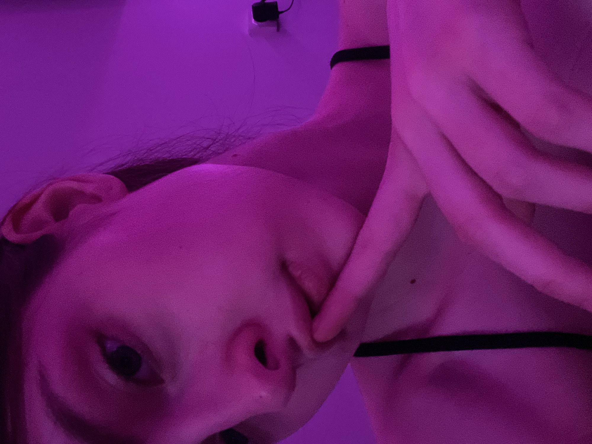 A close-up, low-angle shot of a person's face, lit with a strong purple hue. The individual has light skin, dark hair, and their left eye is visible. Their hand is near their mouth. A black choker-style necklace is visible on their neck. The background is blurred but appears to be a plain wall. A power adapter is mounted high and slightly behind. The location is not discernible based on the image.