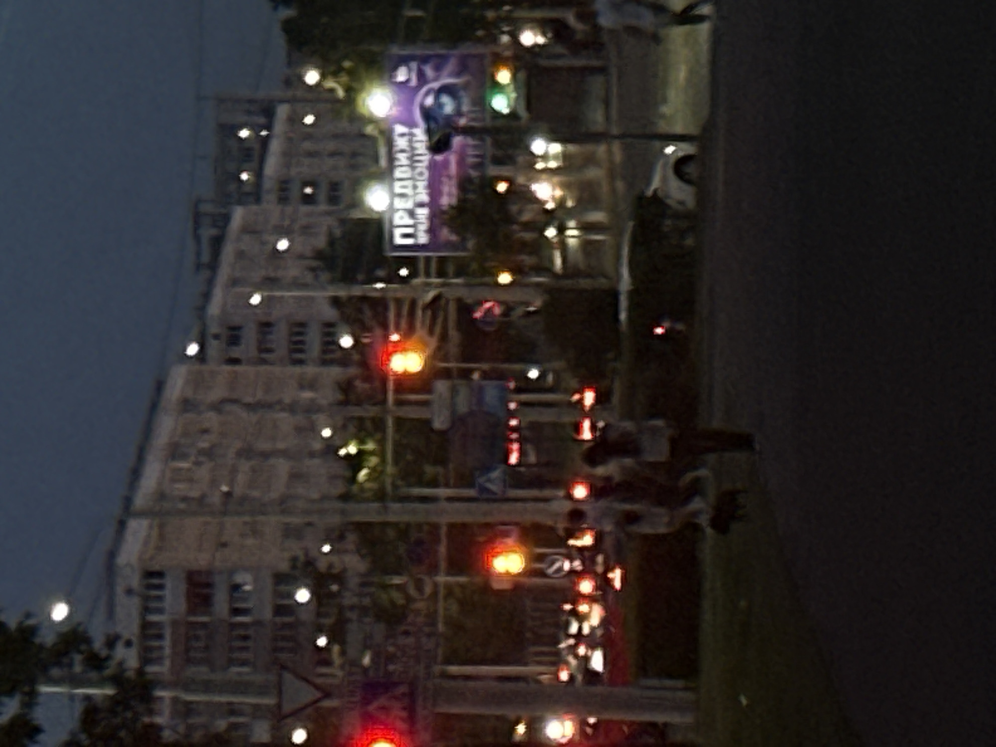 An outdoor, slightly blurry, night-time scene depicts a city street. In the background, a large, multi-story apartment building with numerous windows dominates the skyline. In the mid-ground, a large, illuminated billboard displays text and an image of an eye. Various traffic lights are visible, some red, some orange, and some green, indicating an intersection. Several streetlights cast a glow on the surroundings. In the foreground, a few individuals are walking on a sidewalk. A small, dark-colored dog is also present on the grass. A white car is parked on the side of the road. The overall impression is one of a typical urban environment at dusk or night.