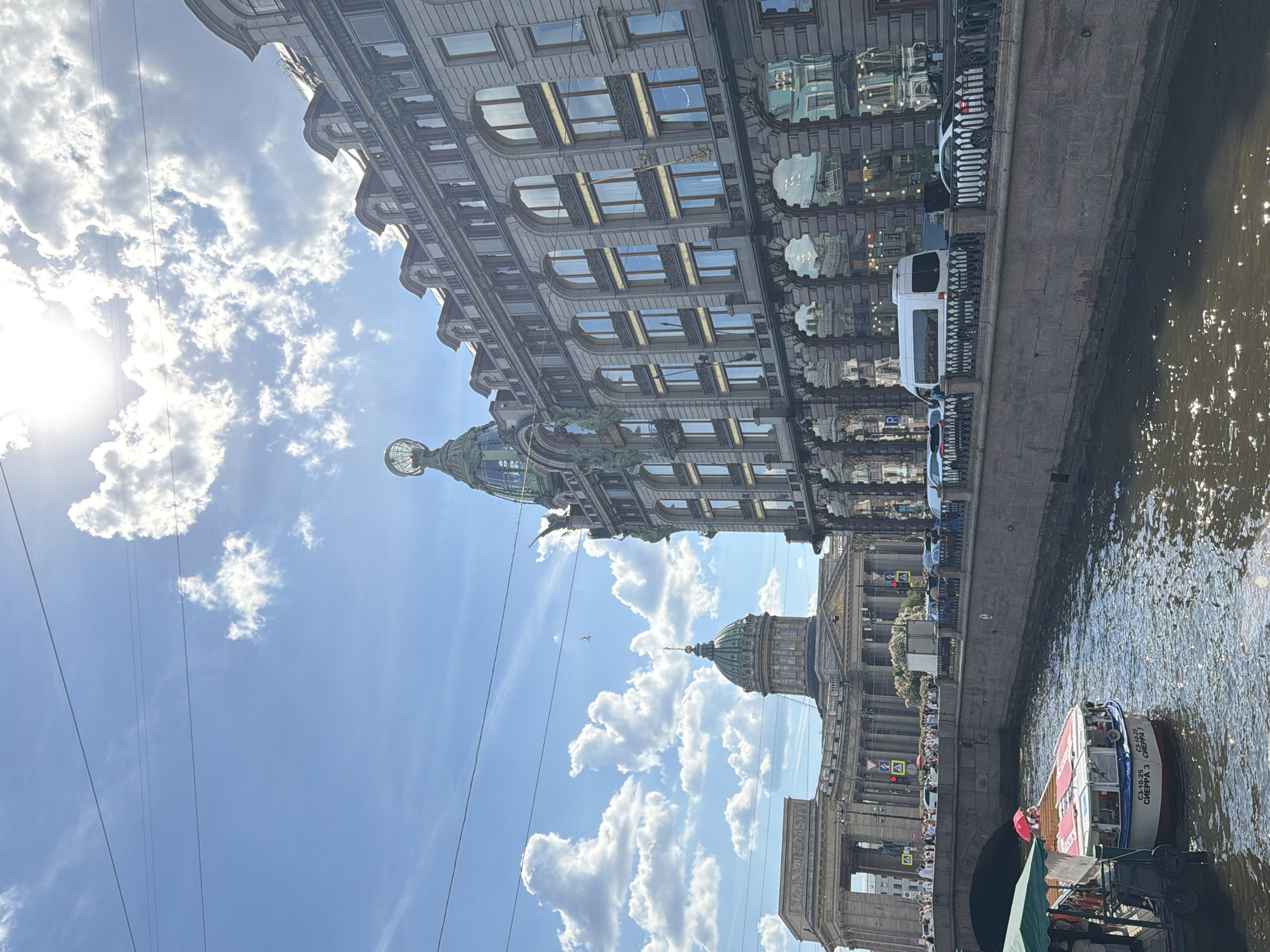 This daytime image captures a scenic view of Saint Petersburg, Russia, centered around a canal, likely the Griboyedov Canal, flanked by prominent architectural landmarks under a bright, sunny sky. The sun shines intensely from the left, illuminating scattered white clouds across the blue expanse, with some faint overhead wires also visible.
On the left side of the canal stands the ornate Singer House (Dom Knigi), a multi-story building characterized by its elaborate architectural details, numerous large windows, and a distinctive glass dome topped with a large globe. Reflected light from its windows suggests a vibrant interior or street scene. On the opposite side of the canal, partially obscured, is the grand Kazan Cathedral, identifiable by its classical columns and a prominent green dome.
A stone embankment with a dark metal railing separates the canal from a street where several cars, including a white van, are visible. The canal's water, dark with ripples, reflects the bright sunlight. A tour boat, light-colored with red accents and an awning, is seen on the canal, carrying passengers. Visible text on the boat includes "СЗ 10-25", "СВЕРХ 3", "СВЕРХ 7", and "ARSENAL".
Numerous people are present on the embankment and on the boat, suggesting a bustling tourist area. A small bird can be seen flying in the clear sky above. The scene conveys a lively atmosphere on a pleasant, clear day in the city.