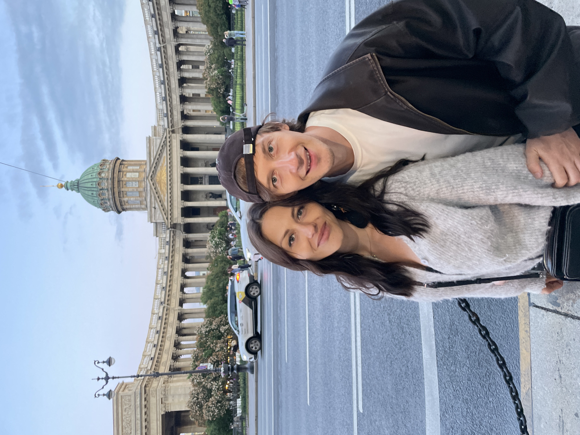 A man and a woman stand in the foreground of the image, posing for a selfie. The woman is on the left, wearing a light gray, fuzzy cardigan over a dark top, and a black crossbody bag. The man is on the right, wearing a white t-shirt and a dark brown leather jacket, with a dark baseball cap. They are positioned on a sidewalk in front of a street with visible lane markings.
Behind them, a large, classical-style building with a prominent green dome and colonnades stretches across the background. This is Kazan Cathedral, a landmark in Saint Petersburg, Russia. The sky above is overcast with a pale blue hue. Trees and more of the building's facade are visible to the sides. A lamppost stands to the left of the couple. A white vehicle, possibly a taxi with yellow markings, is parked on the street. In the middle ground, a decorative chain fence runs along the edge of the sidewalk. Several other people are scattered in the background, some walking and others gathered near the building.