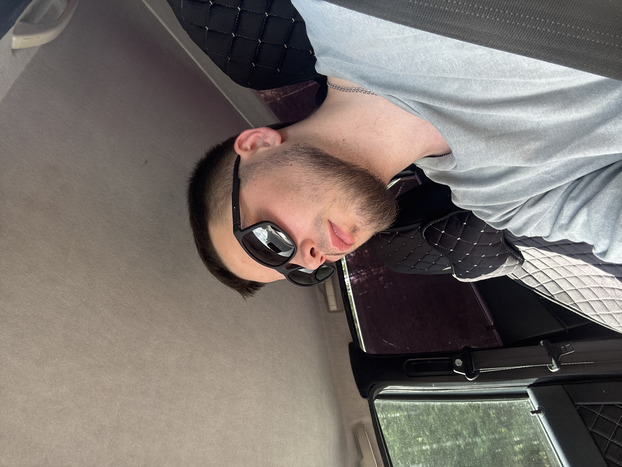A man with a short haircut, beard, and sunglasses is in the passenger seat of a car. He is wearing a light blue t-shirt and a silver chain necklace. The car's interior is visible, including a black seat with diamond stitching, a seatbelt, and a window with trees outside. The scene is set in Verkhnyaya Talitsa, Russia.