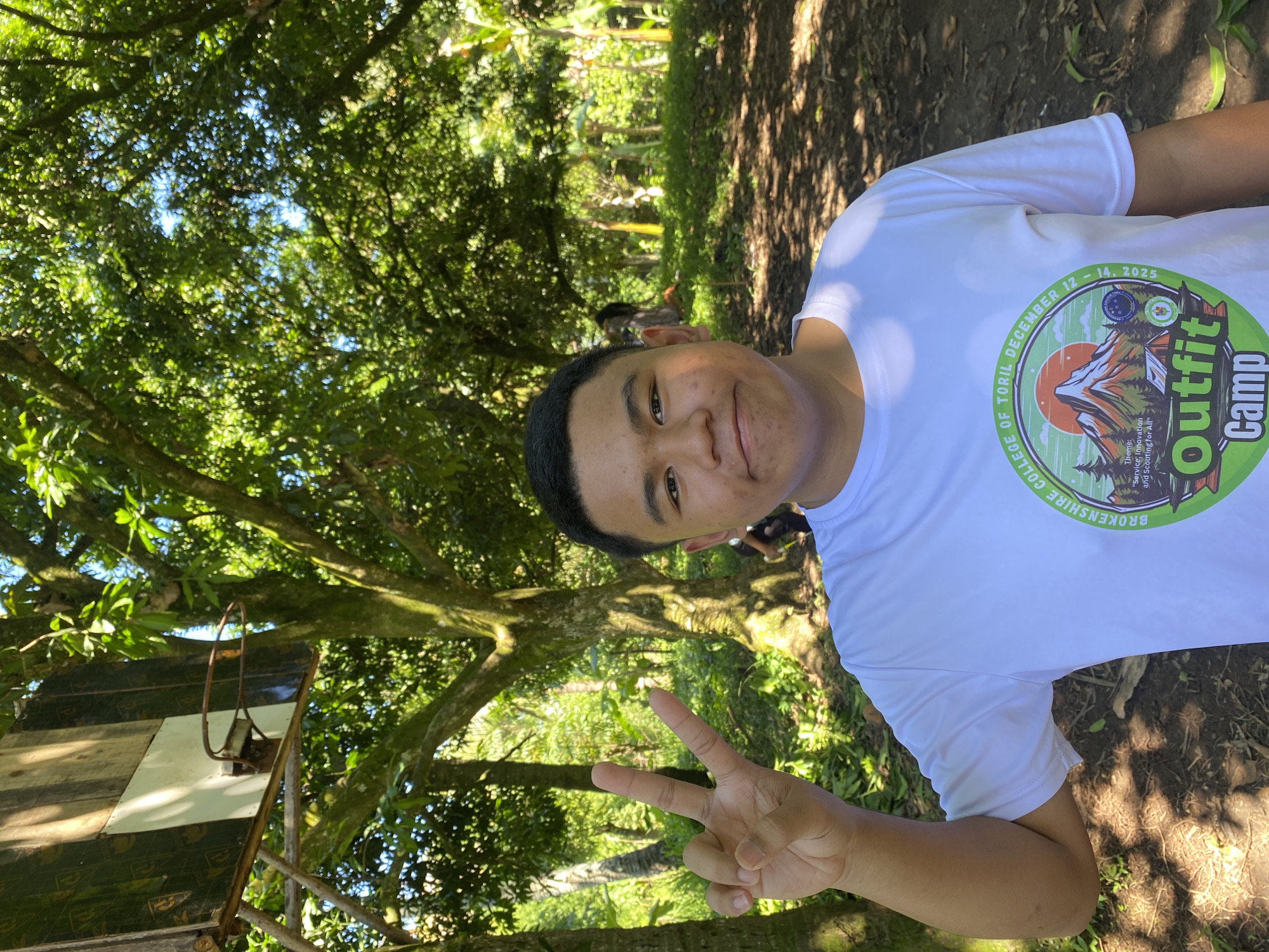 A young male is posing for a selfie outdoors, making a peace sign with his left hand. He is wearing a white t-shirt with a circular logo that says "BROKENSHIRE COLLEGE OF TORIL DECEMBER 12 - 14, 2025" and "Outfit Camp" below a mountainous landscape graphic. He has short dark hair and a light complexion.
The background consists of lush green foliage, including large trees with visible branches and leaves, suggesting a tropical environment. Sunlight filters through the leaves, creating dappled light on the ground, which is covered with dirt and fallen leaves. To the left, a portion of a makeshift basketball hoop and backboard is visible, constructed from wood and metal. In the far background, other trees and vegetation are visible, with a hint of sunlight indicating a clear sky. The overall scene appears to be a park or a natural outdoor recreational area within Davao City, Philippines, likely a campsite or a place associated with the Brokenhire College of Toril.