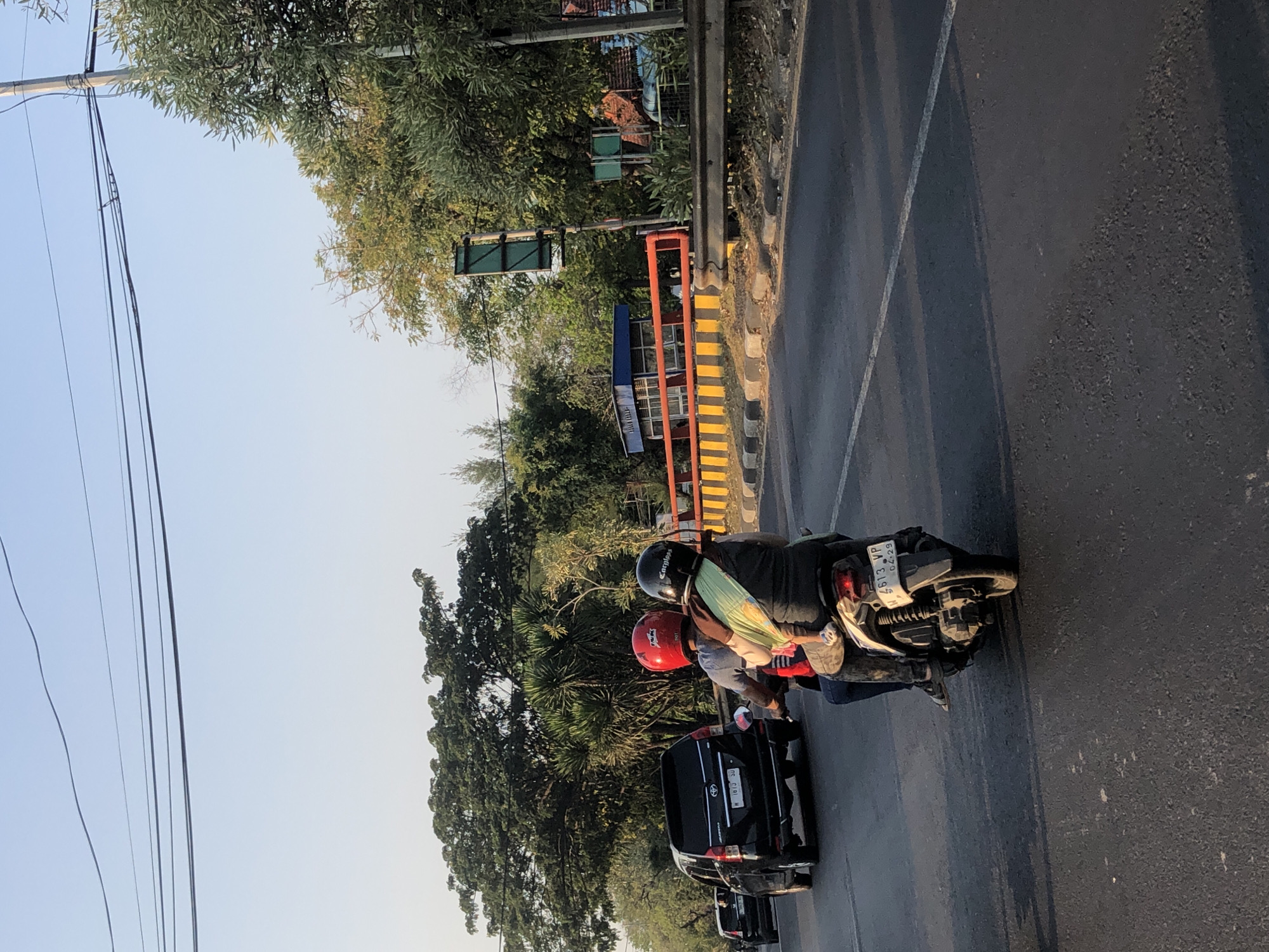 The image depicts a street scene in the city of Ngaban, Indonesia, likely during daylight hours given the clear sky. In the foreground, two people are riding a scooter. The person in the back, presumed to be the driver, wears a red helmet. The passenger, seated behind the driver, wears a black helmet and is holding a young child who is also wearing a helmet. The passenger has their arms around the child. The scooter is white and has a license plate that reads "W 4613 VP 04-29".
To the left of the scooter, a black SUV is driving in the same direction. The SUV has a license plate that reads "W 1813 SU". Further in the background, more vehicles are visible, suggesting moderate traffic.
The environment is characterized by a paved road with a white line marking a lane. On the right side of the road, there is a guardrail and a sidewalk area. Beyond the sidewalk, there are various trees and foliage, with some buildings and structures visible in the distance, including a blue and orange structure that appears to be a bus stop or a small shelter. Utility poles and power lines are also present in the scene, crossing the sky. The overall impression is a typical urban or suburban street in Indonesia.