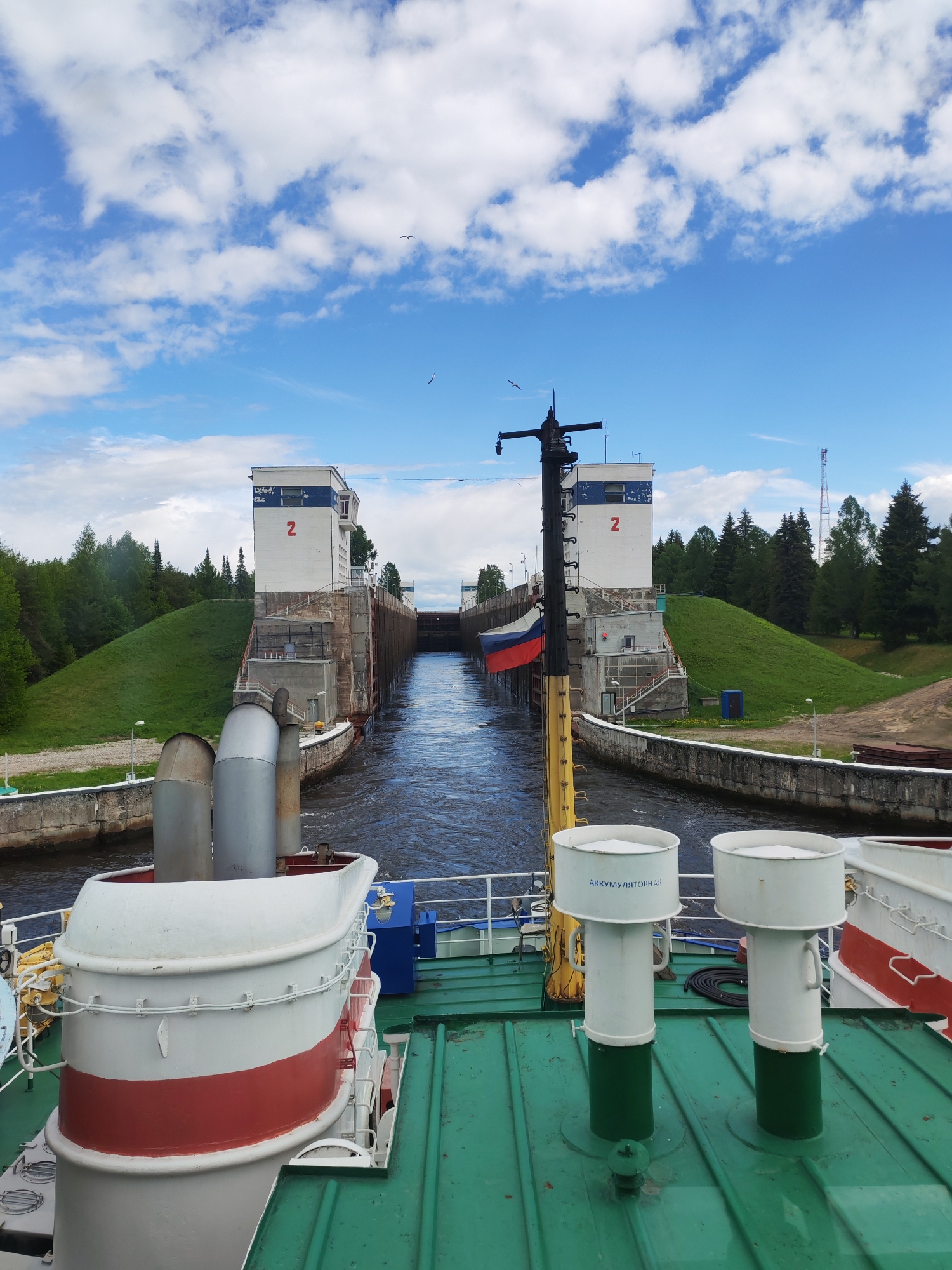 A ship is traversing the Volga–Baltic Waterway in Анхимово, Russia, approaching Lock Number 2. The lock is flanked by identical white buildings, each marked with a large red "2". A Russian flag is visible on the vessel. The sky is bright blue with scattered cumulus clouds. Green grassy slopes and trees border the waterway. The foreground shows the ship's deck, featuring white and red structures, green deck paint, and two white vents with green bases.