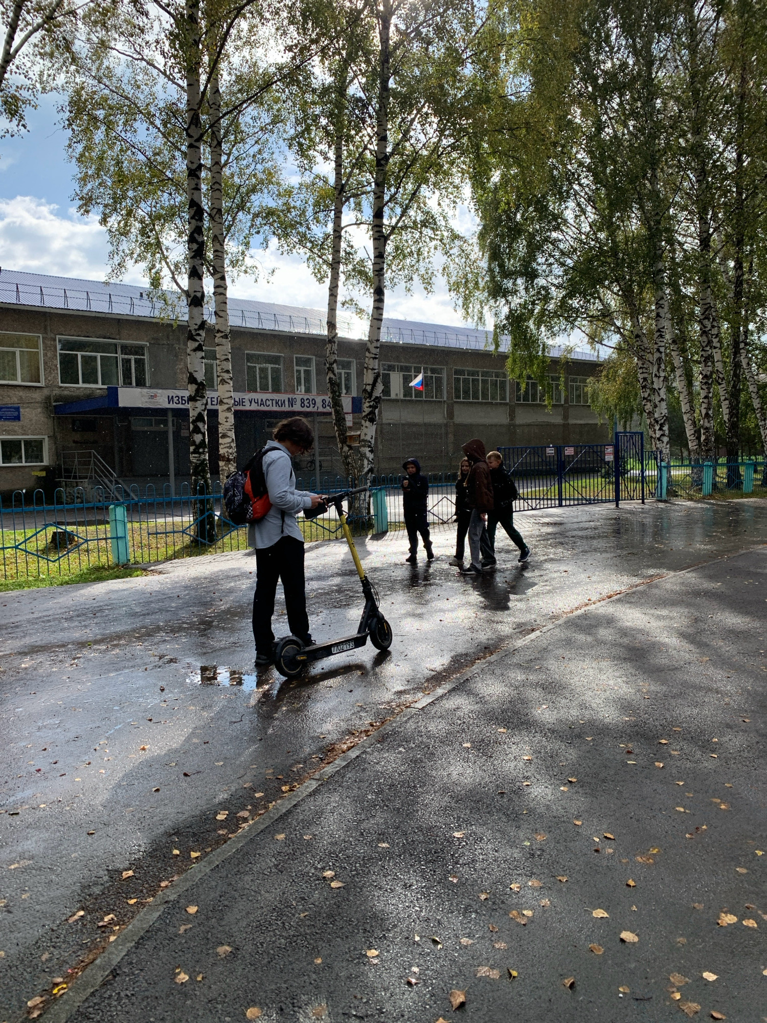 The image depicts the exterior of a school building in Prokopyevsk, Russia, on a wet, overcast day, possibly in autumn, given the scattered fallen leaves on the asphalt. The school building is a typical Soviet-era structure, constructed from concrete blocks with a row of evenly spaced windows across its facade. A sign above the entrance reads, "ИЗБИРАТЕЛЬНЫЕ УЧАСТКИ № 839, 840" (Election Precincts No. 839, 840). A Russian flag is visible on the building.
In the foreground, a young person with dark, curly hair wearing a light blue shirt and dark pants stands on a wet asphalt path, looking at their phone. They have a backpack with red and black accents. Beside them is a black electric scooter with yellow accents. The scooter has a visible license plate with the characters "7 JO249". Puddles of water are present on the ground, reflecting the surroundings.
To the right of the main subject, a group of four young people are walking away from the camera, towards the school entrance. They appear to be students. One person in the foreground is wearing a dark hooded jacket, followed by two individuals in dark clothing, and another person in a brown jacket.
The area is bordered by a blue metal fence. Several birch trees with distinctive black and white bark line the background, their leaves appearing green but somewhat sparse, suggesting the season. The overall atmosphere is damp and subdued, with sunlight occasionally breaking through the clouds.