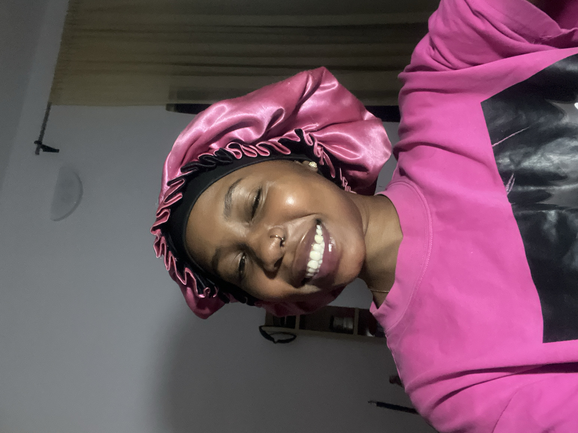 A young Black woman with dark skin is smiling widely and looking at the camera in a close-up shot. She is wearing a bright pink long-sleeved sweatshirt with a black graphic design on the front and a shiny pink satin bonnet with a black band. She has a nose ring. The background is dimly lit with a gray wall and beige curtains. The setting appears to be indoors, possibly a bedroom, and the lighting suggests it is nighttime or a dimly lit room. There are no indications of the location being specifically in the city of Jos, Nigeria.