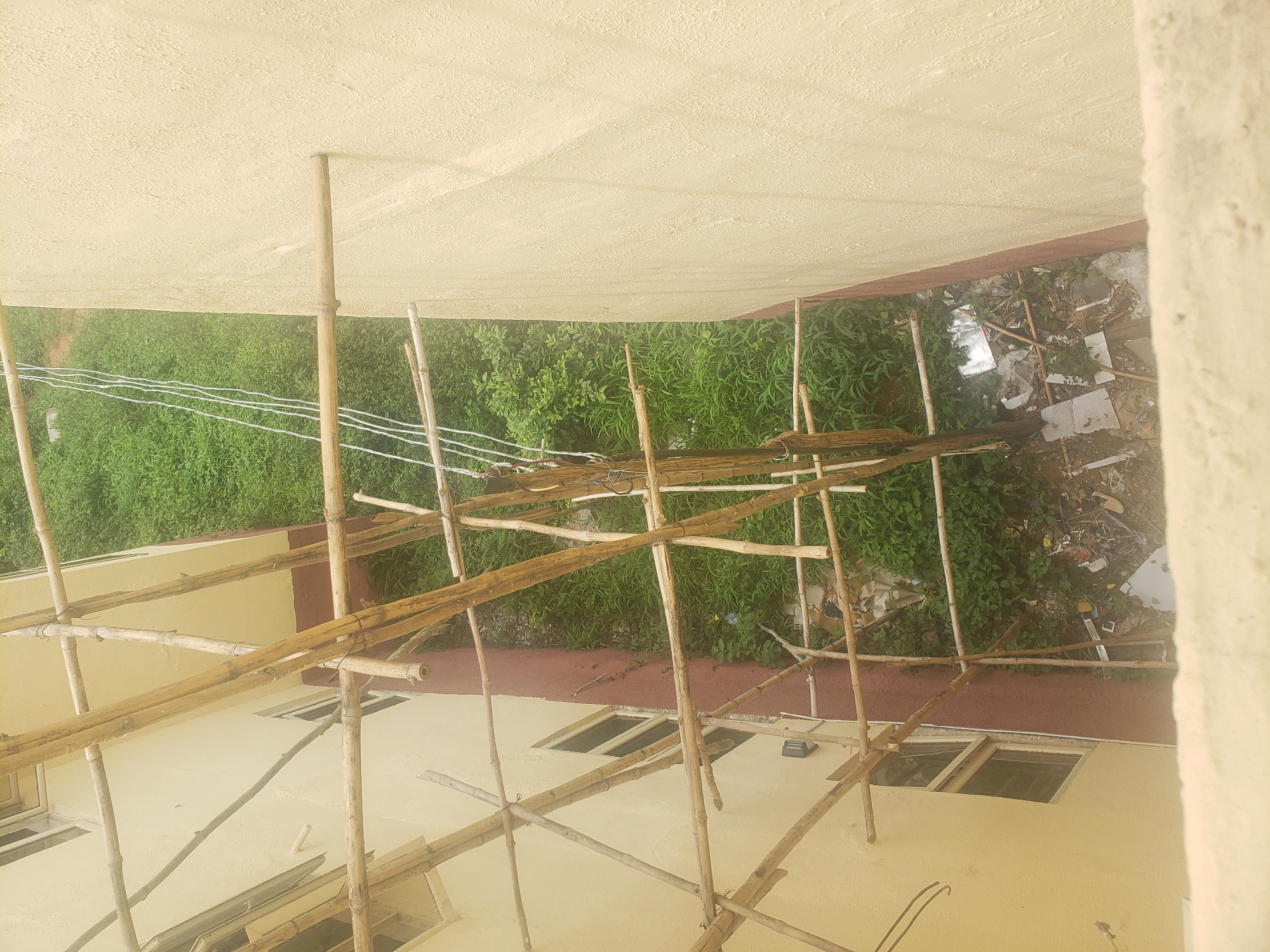 The image captures a scene from an elevated perspective, looking down at the side of a building under construction or renovation. Bamboo scaffolding is erected along the building's exterior, which has a light yellow, textured facade. To the left of the building, windows with metal grates are visible. Between the building and a dense patch of green vegetation, a narrow strip of ground is littered with debris, including plastic bags, paper, and wood scraps. Several white ropes are strung vertically in the upper right portion of the frame. The overall impression is one of an unfinished outdoor area adjacent to a building, likely in a tropical or sub-tropical environment given the lush greenery. The location is identified as Gumel, Nigeria.