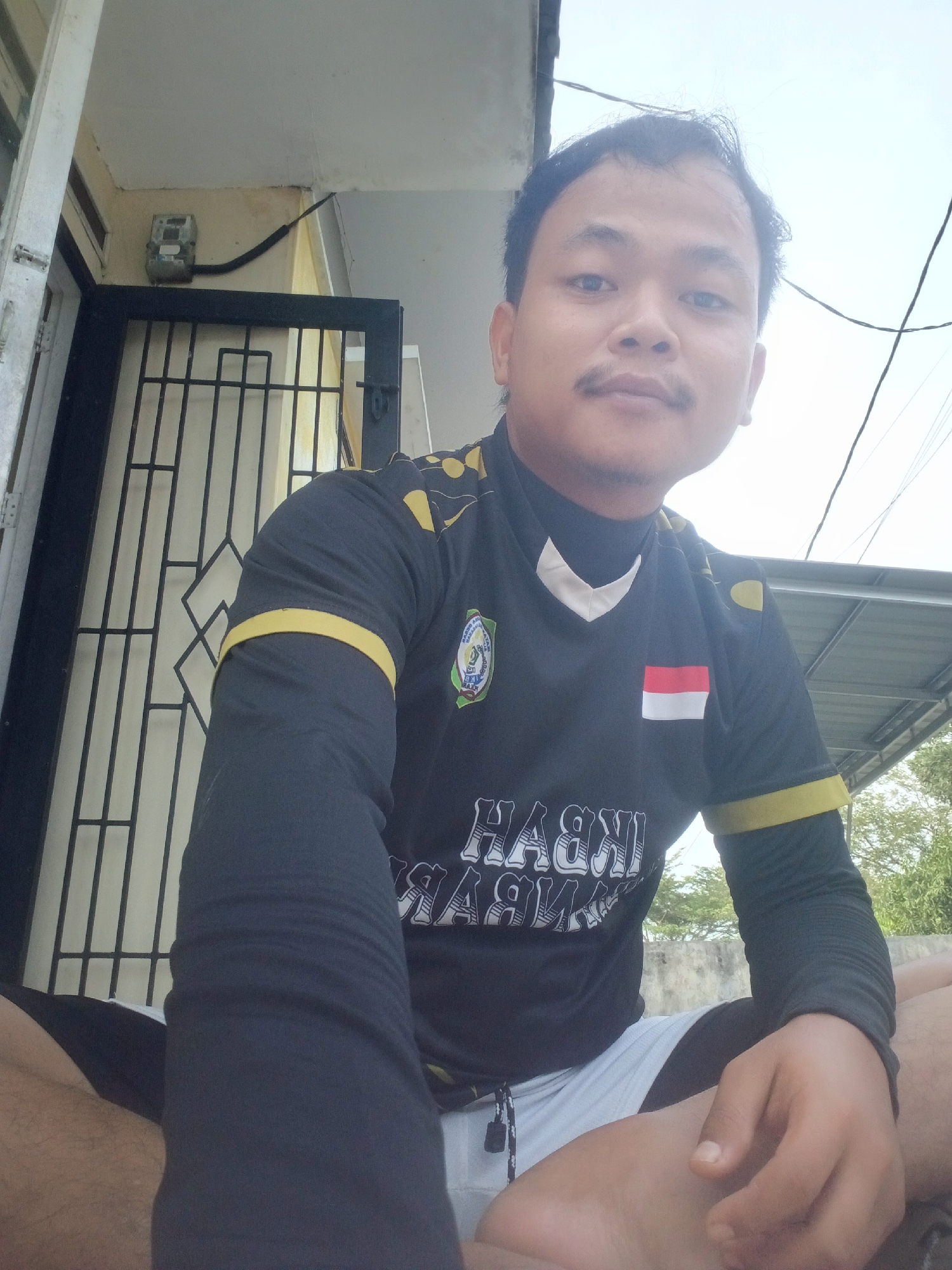 In Kualu, Indonesia, a young man is photographed from a low angle, looking directly at the camera. He is wearing a black long-sleeved shirt with yellow trim on the collar and sleeves. A small Indonesian flag is visible on the right breast of his shirt, and a circular emblem is on the left. The shirt also features white lettering across the chest. He appears to be wearing light-colored shorts over black underwear. The man is seated, with his legs crossed, and his hands resting on his legs. The background shows a building with a metal gate and an electric meter on the wall. A portion of a metal roof structure is visible above and to the right of the man, with trees and a cloudy sky in the distance. The overall lighting suggests it is daytime.