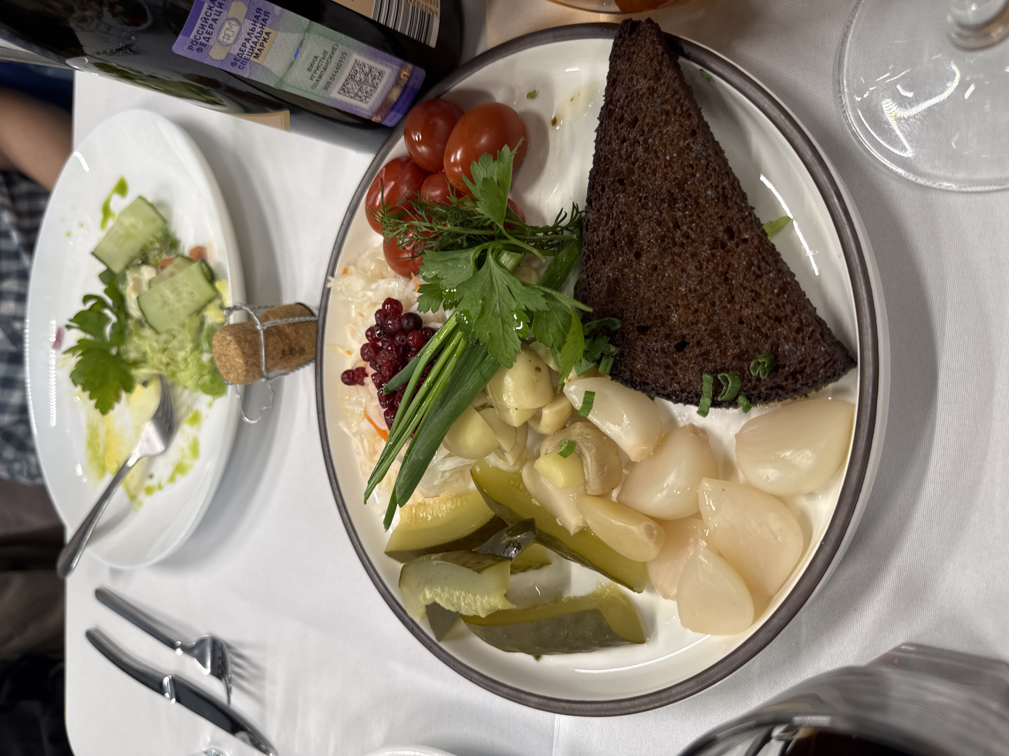 A high-angle shot reveals a dining table setting in Nizhny Novgorod, Russia. A white plate with a dark rim is centrally placed, holding an assortment of food items. These include dark rye bread cut in a triangular shape, pickled onions, sliced pickles, cherry tomatoes, a sprig of dill, parsley, spring onions, sauerkraut, and red berries. To the left, a smaller white plate contains a partially consumed salad with cucumber and a dressing. Next to it, a fork rests on the plate, alongside the cork from a bottle of champagne.
The edge of a bottle with a Russian Federation tax stamp is visible at the top. Cutlery is placed to the lower left of the frame. A partially visible glass and bottle suggest more people may be dining at the table. The tablecloth is white, creating a clean backdrop for the meal.