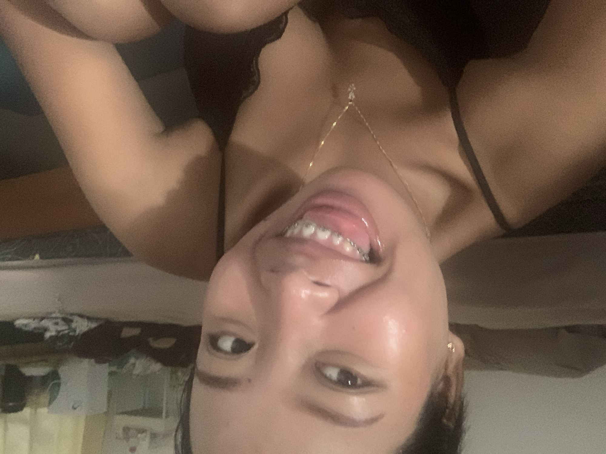 A woman with dark hair and brown eyes is shown from a low-angle, inverted perspective. She has braces on her upper teeth and is wearing a black lace top with thin straps. A delicate gold necklace is visible. Her skin is fair and appears to be well-lit. The background is blurred, but a shelf with various objects can be seen. There is no indication of the location being Denpasar, Indonesia.