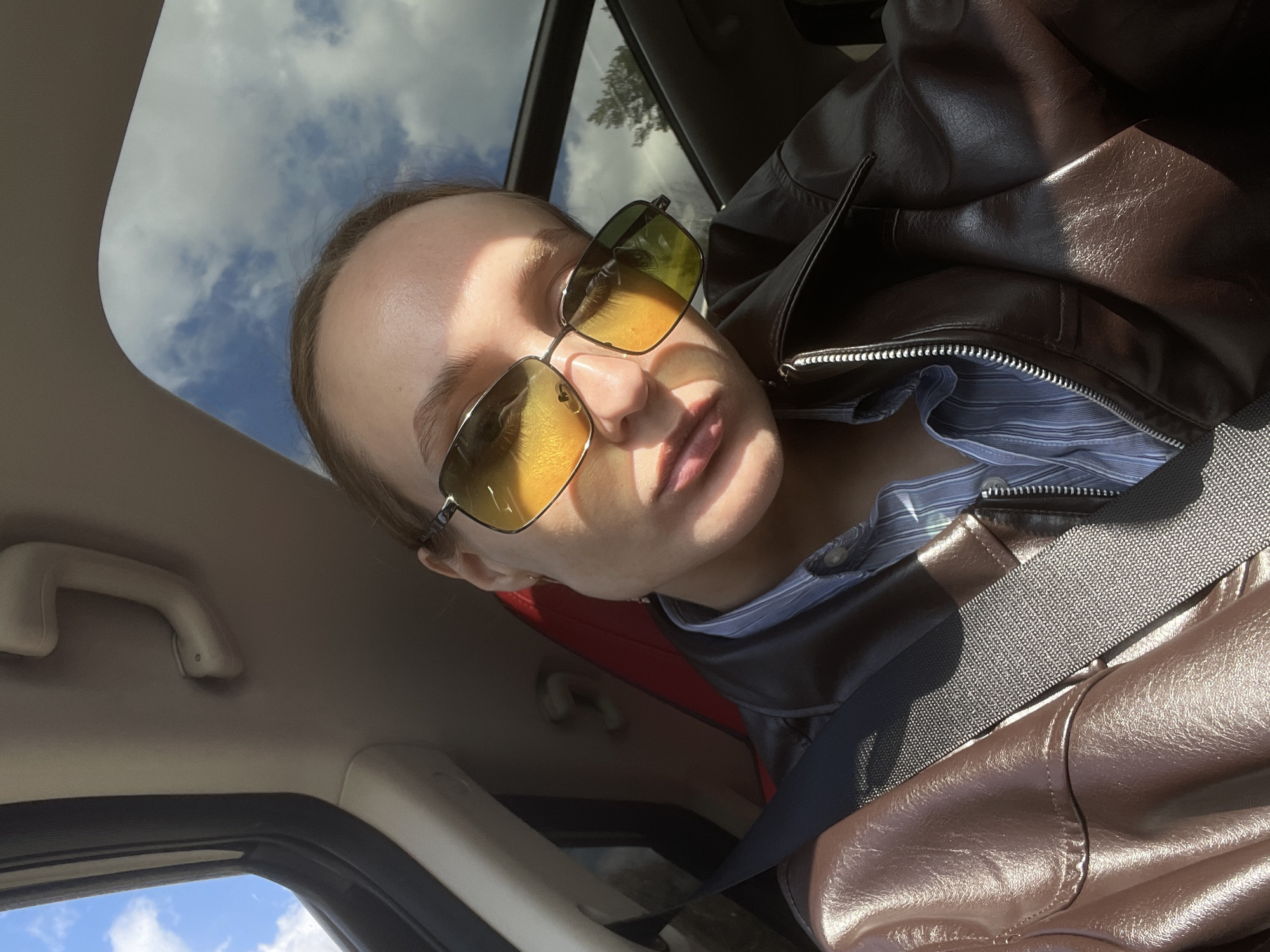 The image is a tilted, close-up shot taken from the interior of a car, featuring a person wearing sunglasses. The perspective looks slightly upwards and outwards through a sunroof and side window.
The main subject is a person, likely in the driver's or passenger's seat. Their face, from the forehead to the chin, is visible, obscured by large, aviator-style sunglasses with yellow-orange tinted lenses. The person's hair is pulled back. They are wearing a dark brown, possibly leather, jacket with a visible zipper detail, over a blue and white striped shirt. A dark grey seatbelt crosses diagonally across their chest and jacket.
The setting is the interior of a vehicle. Above the person's head, a car's ceiling and a beige grab handle are visible. Through the car's sunroof and a side window on the left, a clear blue sky with scattered white clouds is seen. The interior of the car also shows a dark window frame and hints of a red surface behind the person's shoulder, possibly part of a car seat or interior trim.
The activity appears to be posing for a photo while in transit or parked. The time of day is daytime, with bright, direct sunlight casting strong highlights on the person's face, sunglasses, and jacket, as well as creating distinct shadows. The weather is sunny with some clouds. No specific emotions are discernible due to the sunglasses and lighting. No text is visible in the image. The location context provided is Ol'khovochka, Russia.