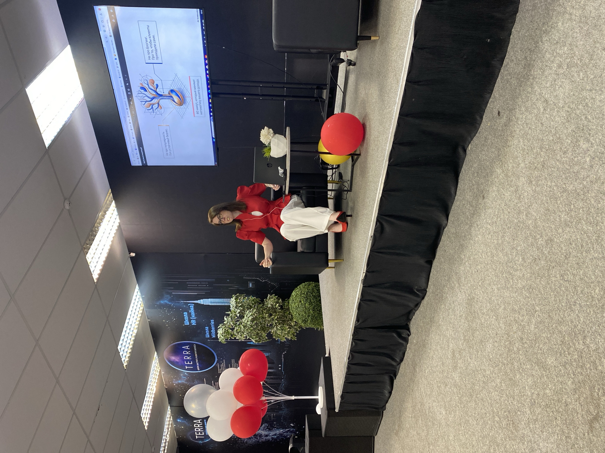 The image depicts an indoor event space. A woman in a red blazer and white pants is seated on a black sofa on a raised stage. She is looking towards a large screen displaying a presentation with text and graphics. To her left, a laptop is open on a small table, alongside a decorative plant and a white flower. A cluster of red and white balloons stands to the far left of the stage, and two large spherical balloons, one red and one yellow, are on the stage floor in front of the sofa. The stage is covered in gray carpet and is draped with a black skirt. In the background, a black wall features logos and text, including "TERRA" and "Школа HR (наймa)." The ceiling has fluorescent lighting panels. The overall setting suggests a conference, seminar, or presentation event.