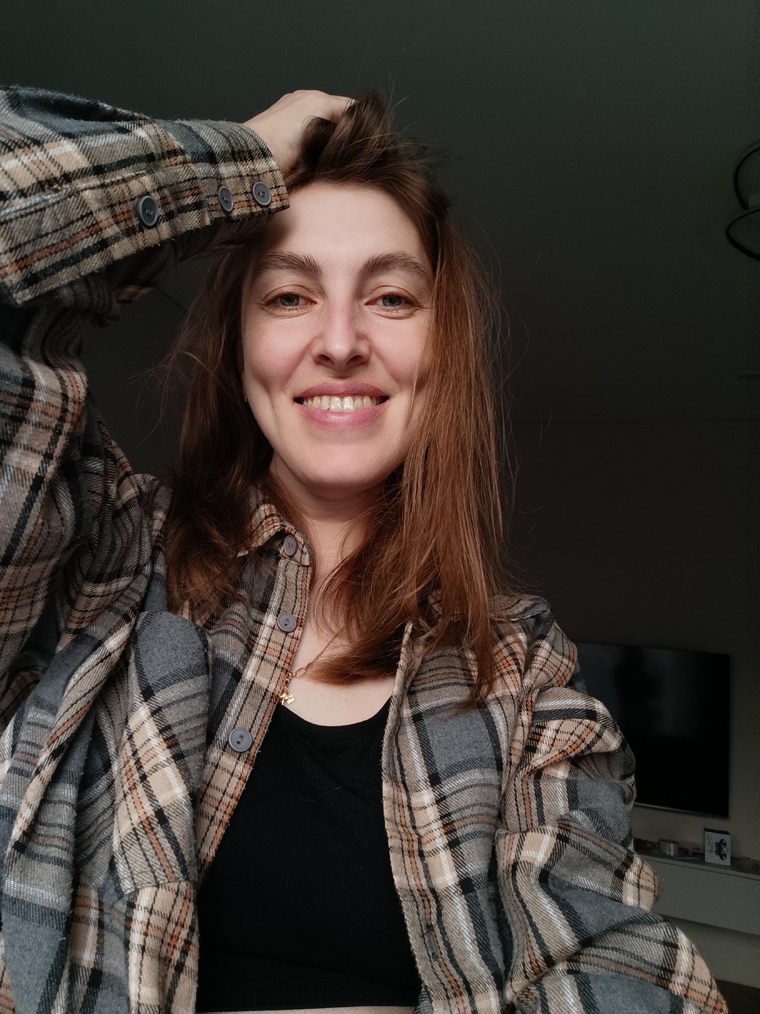 In the provided image, a woman with medium-length brown hair is captured in a close-up shot, looking directly at the camera with a wide smile that reveals her teeth and dimples. She is wearing a plaid shirt over a black top and has her left hand raised, with fingers tangled in her hair. The background is dimly lit, suggesting an indoor setting, possibly a home in Chaikovsky, Russia, given the context. The lighting is soft, with the woman's face illuminated by a light source from the front. The image appears to be a selfie, taken at a slightly low angle.