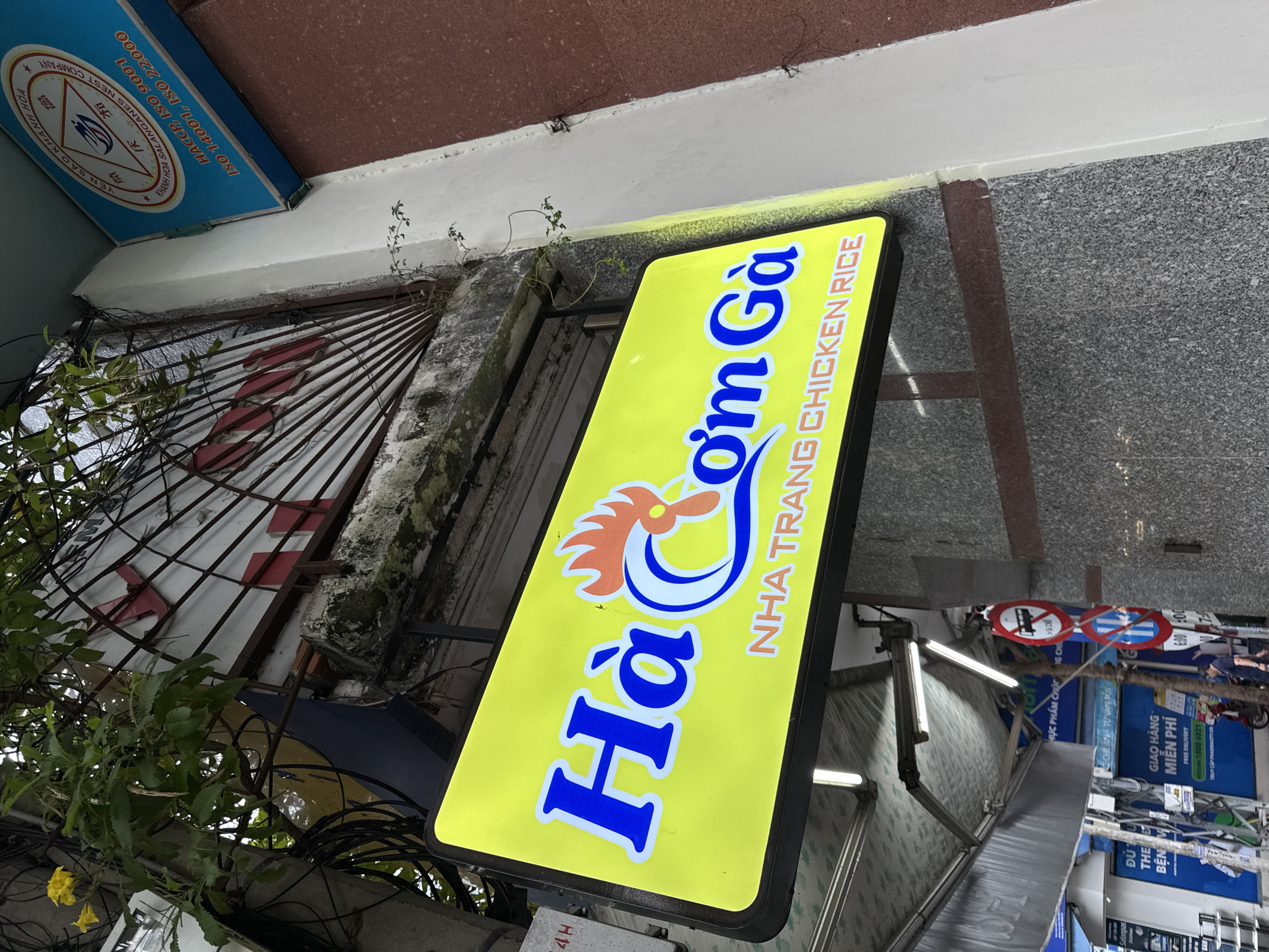 A street-level, eye-level shot captures a neon sign advertising "Hà COM GÀ NHA TRANG CHICKEN RICE". The sign is predominantly yellow with blue and orange lettering, featuring a stylized image of a chicken. Below and to the right of the sign, a light-colored building wall is visible, with a darker, granite-like wall at the lower right. To the left of the sign, a dense network of electrical wires hangs in front of a metal grate. In the background, an awning with fluorescent lights is visible, and beyond that, a building with blue signage. Several street signs and a motorbike are also partially visible in the background, suggesting an urban environment in Nha Trang, Vietnam.