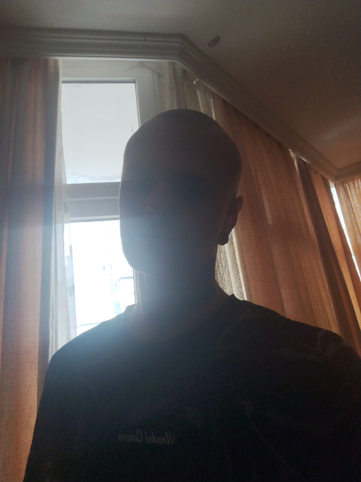 The image shows a person's head and shoulders against a window with light filtering through curtains. The person is bald and appears to be in silhouette due to the strong backlight from the window. The environment suggests an indoor setting, possibly a residential room, with curtains on either side of the window and crown molding along the ceiling. There is a faint inscription visible on the person's shirt, but it is illegible due to the low light conditions and blurriness. The scene does not provide any specific details to pinpoint its location within the city of Batumi, Georgia.