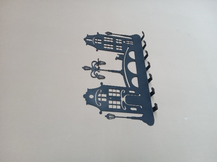 A decorative metal wall-mounted coat rack, powder-coated in a dark blue-grey. It's shaped like Dutch canal houses with cut-out window details, flanked by lampposts. A curved section runs down the side, punctuated with half-circles and ending in individual hooks for hanging items. The rack is mounted against a plain, off-white wall.