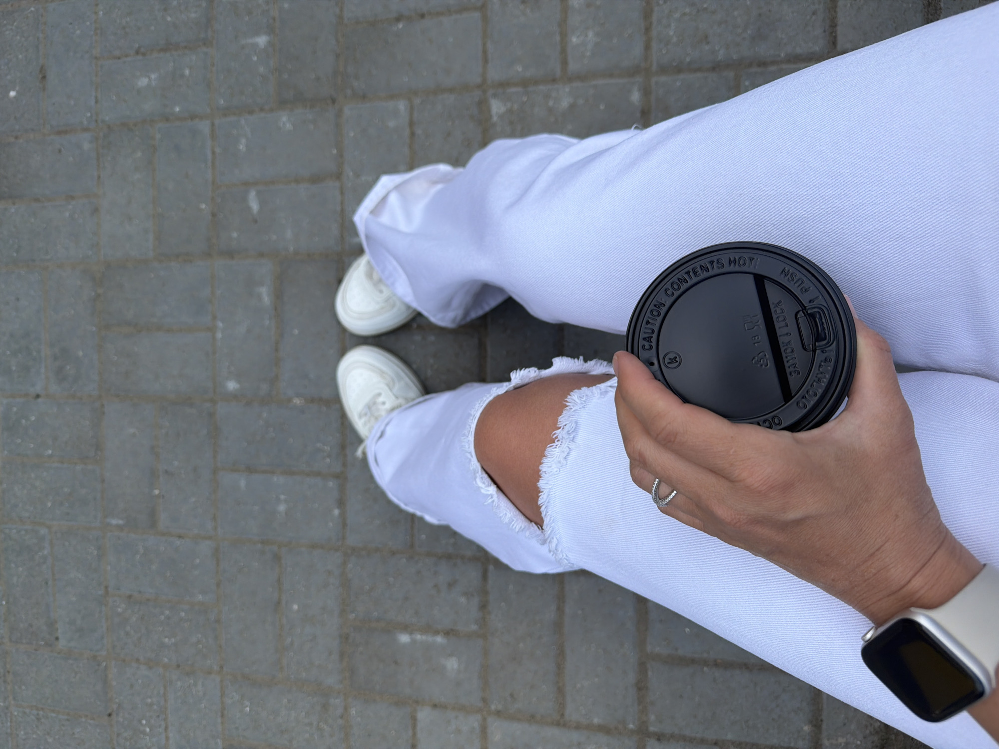 A top-down shot depicts a person seated on gray brick pavers, presumably in Chaikovsky, Russia. The person wears white jeans with a frayed hole at the knee and white sneakers. They hold a black disposable coffee cup lid in their right hand. A silver ring is visible on the ring finger of their right hand. A light-colored smartwatch is on their left wrist.