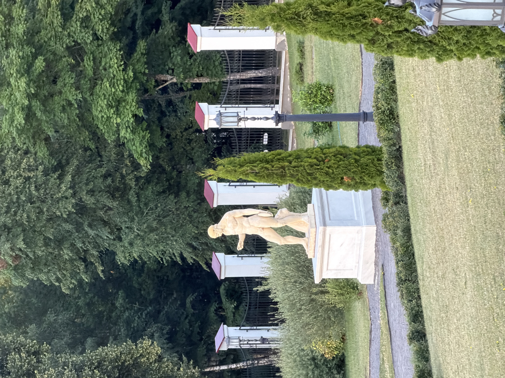 The image depicts a classical statue of a discus thrower on a pedestal, positioned in an outdoor garden setting. The statue is rendered in a light-colored material, possibly marble or stone, and appears to be in motion. It is surrounded by lush green trees and manicured bushes. A paved pathway runs alongside the grass, leading further into the garden.
In the background, there are several white structures with red roofs, resembling ornate lampposts or decorative architectural elements. Black wrought-iron fencing is visible behind these structures. The overall atmosphere is serene and suggests a well-maintained park or historical garden. The lighting indicates it might be late afternoon or early evening, with soft shadows present. The weather appears to be clear, contributing to the pleasant outdoor ambiance. There is no visible text in the image.