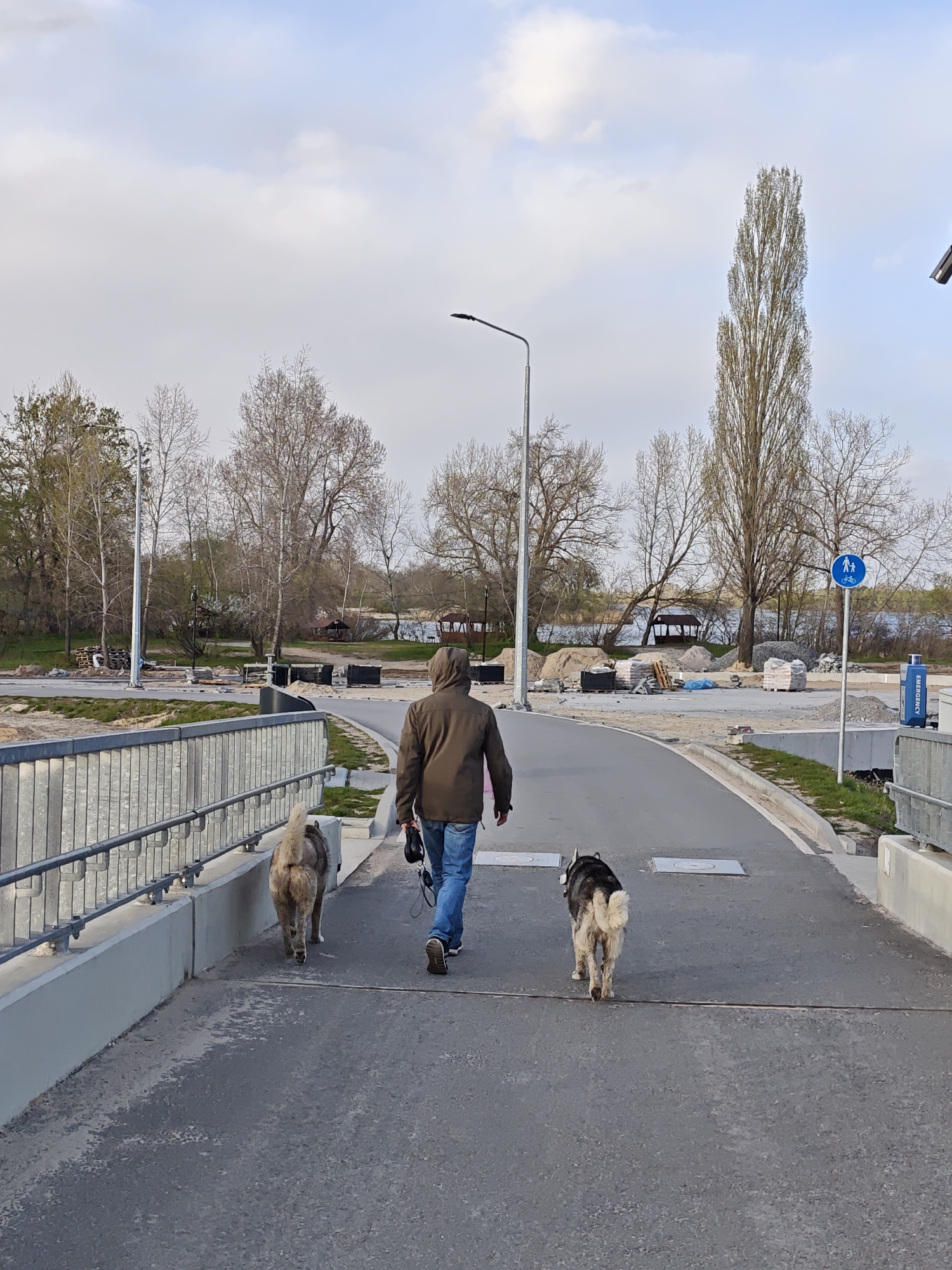 A person is seen from behind, walking two dogs on a paved path or bridge. The individual wears a brown hooded jacket and blue jeans, holding leashes in their left hand. Two large dogs accompany them; one light-colored and fluffy walks near the left railing, while the other, with dark fur and a lighter, fluffy tail, walks slightly to the right.
The path, which appears to be part of a pedestrian or cycling route, curves gently to the right. The left side of the path features a sturdy metal railing overlooking what appears to be a lower level or waterway. Beyond the curve, the scene opens to an area showing signs of ongoing construction or landscaping work, with piles of sand, stacked bricks on pallets, and black construction containers. In the background, numerous trees, many still bare or just beginning to leaf out, line the edge of a body of water, likely a river or lake, characteristic of a park or riverside area in Kyiv, Ukraine. A prominent, tall, slender tree stands on the right, alongside a street lamp.
A blue circular sign on the right indicates a shared path for both bicycles and pedestrians. Further right, a blue box labeled "EMERGENCY" is visible. The sky above is partly cloudy with patches of blue, suggesting a mild daytime, possibly spring, setting. The overall impression is a casual walk through a developing urban green space.