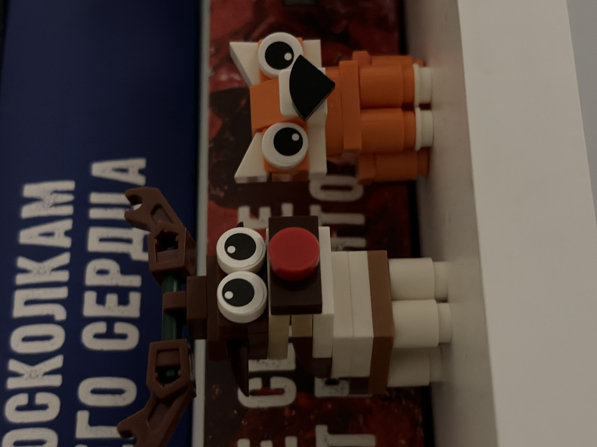 The image shows two small Lego-style figures, a fox and a reindeer, placed on top of books or magazines. The fox is orange and white with a black nose and large eyes. The reindeer is brown and white with antlers and a red nose. The books/magazines underneath have text in Russian. The location is unspecified and cannot be confirmed to be in Perm, Russia.