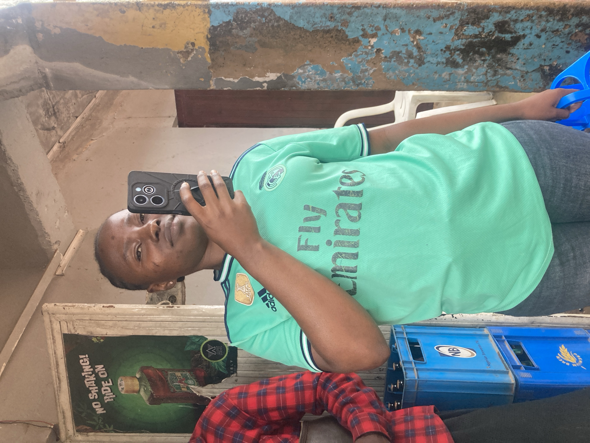 An individual with short dark hair and a medium complexion is captured in what appears to be a selfie or video call, holding a black smartphone with a textured case. They are wearing a light green t-shirt with "Fly Emirates" text and an Adidas logo, paired with dark grey trousers. Their expression is neutral to slightly pleasant as they look towards the camera.
The setting suggests a casual, possibly indoor or semi-outdoor space. The walls behind them feature peeling paint in shades of blue, yellow, and grey, indicative of an older or weathered building. In the background, to the right, stacks of blue crates are visible. One crate clearly displays "NB" and "Nigerian Breweries Plc", while another has "PREMIUM BREW" and a wheat emblem, identifying them as beverage crates. A white plastic chair is also partially visible behind the subject.
To the left of the main subject, a framed poster or sign is affixed to the wall. This poster features a bottle with text that reads "NO SHAKING!", "RIDE ON", "BITTERS", and an "ACE" logo. Part of a second person, wearing a red and black plaid shirt, is visible in the lower right corner of the frame. The presence of Nigerian Breweries crates and the general environment suggest a local establishment in Nigeria, consistent with the provided location context of Jos, Nigeria. The time of day and weather are not discernible.