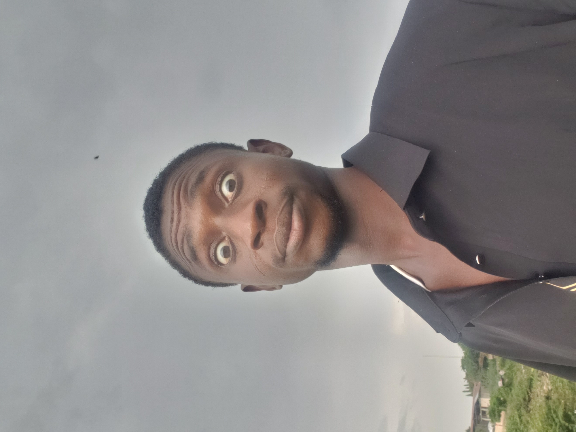 This image is a close-up outdoor portrait of a young man with dark skin, likely taken in Lokoja, Nigeria. He is pictured from the chest up, with his head slightly tilted, and is looking directly at the camera with wide, expressive eyes, conveying an emotion of surprise or intense focus. He has short, dark hair and visible stubble or a short beard. The man is wearing a dark, collared shirt, possibly black, with the top buttons unfastened.
The background consists primarily of a uniform, light grey, overcast sky, suggesting cloudy weather during the daytime. Below the horizon line in the lower part of the image, there are hints of green foliage and some indistinct, distant structures, indicating a semi-urban or rural setting. A small, dark speck, possibly an insect, is visible against the sky in the upper left quadrant. There is no visible text in the image.