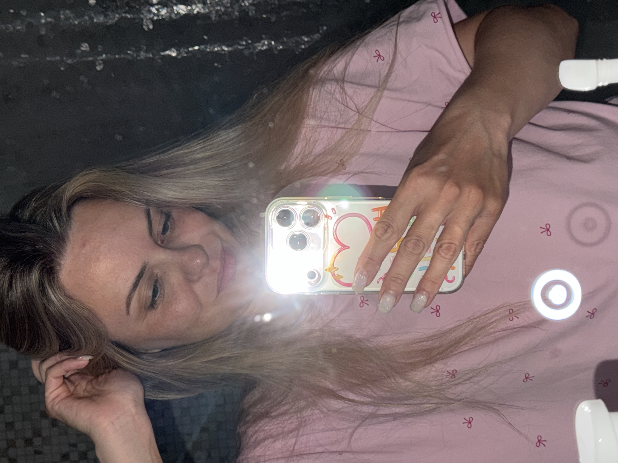 A fair-skinned woman with blonde hair is holding a mobile phone with a heart-shaped decorative case to take a selfie. She is wearing a pink shirt with a small red bow design, and her nails are painted with a glittery finish. The location is implied to be a bathroom as the background shows the reflection of a tiled wall. The environment appears dimly lit with the bright phone camera flash dominating the scene. There is no indication of the image being taken in Votkinsk, Russia.