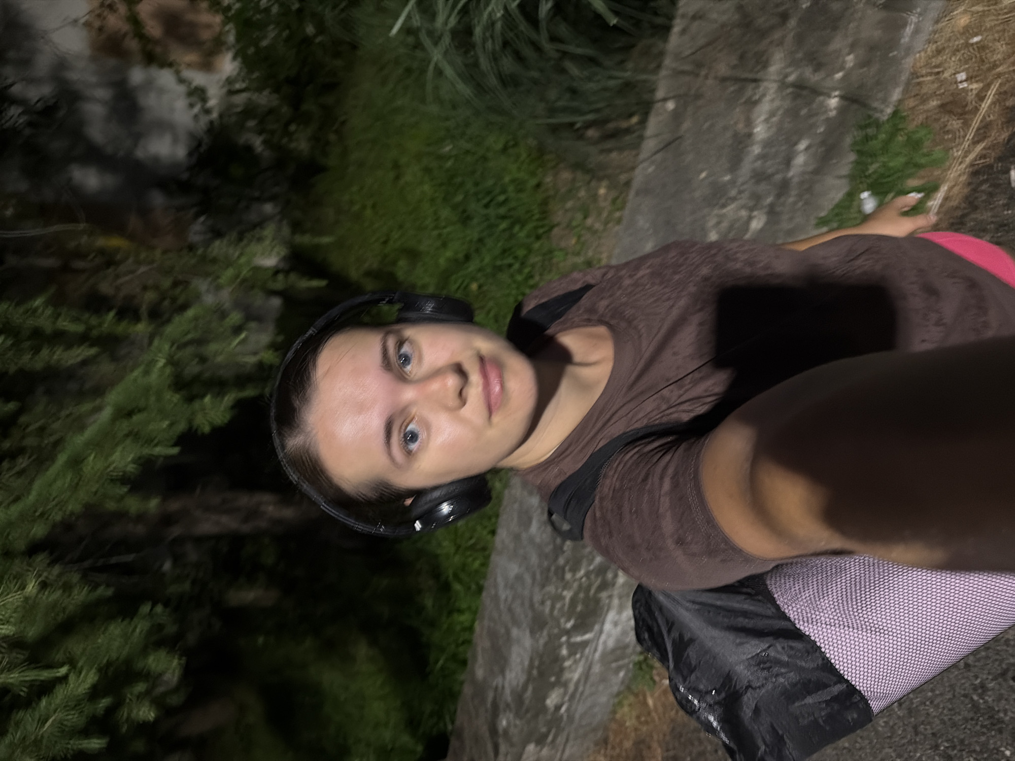 A young woman with blue eyes and light brown hair is taking a selfie in what appears to be a park at night. She is wearing a brown t-shirt, pink shorts, and black headphones. She is carrying a black backpack and a rolled-up purple mat. The background consists of lush green foliage and a concrete structure. The lighting suggests artificial illumination. The setting is consistent with the Arboretum Botanical Garden in Sochi, Russia, known for its diverse flora and nighttime ambiance.