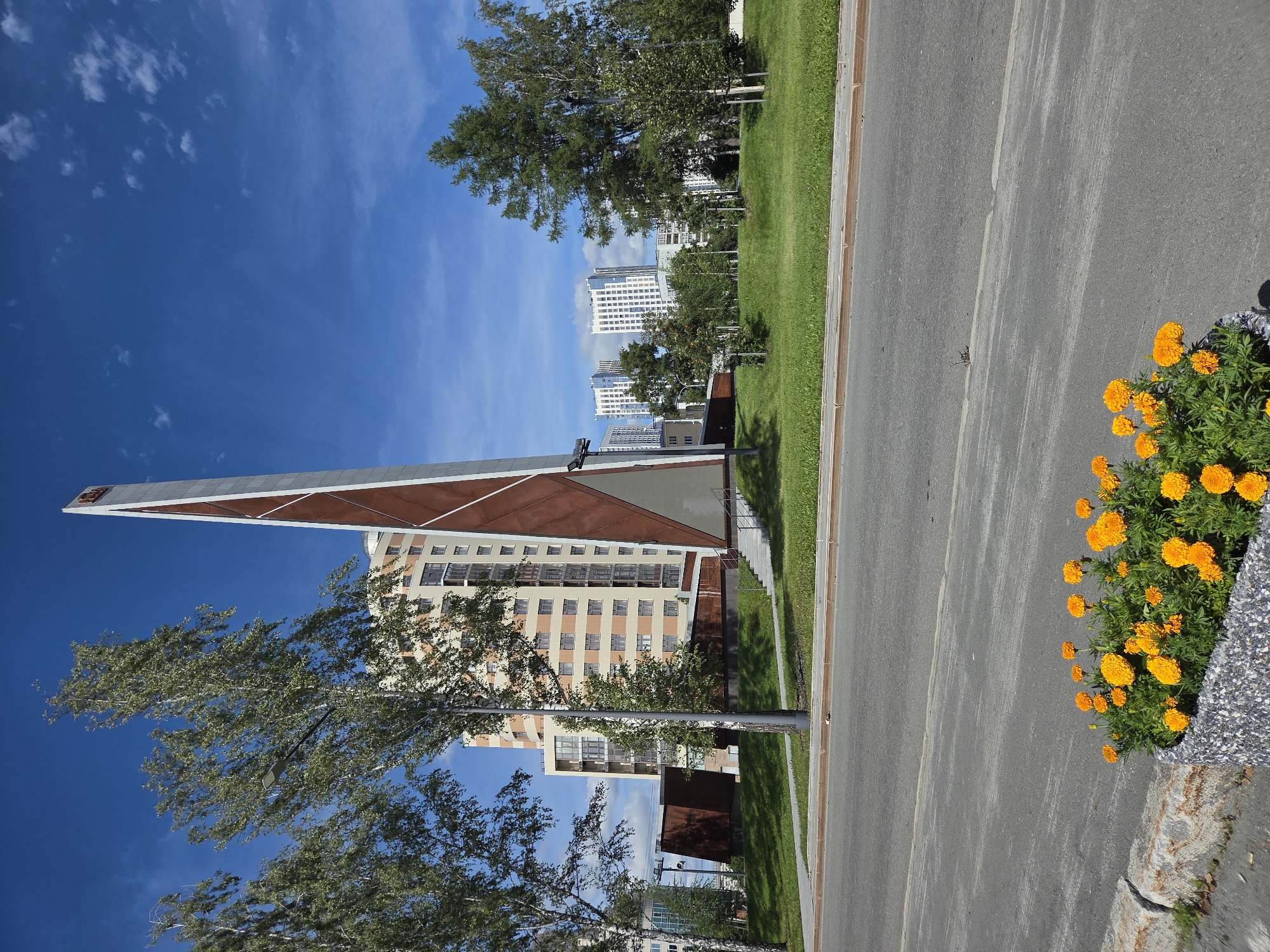 The image depicts a modern urban scene in Yekaterinburg, Russia, under a bright blue sky with scattered clouds. In the foreground, a gray asphalt road runs horizontally across the frame. To the left, a planter filled with vibrant orange marigolds is visible along the curb. A stone border separates the road from a neatly manicured grassy area.
Dominating the midground is a tall, slender, angular monument, primarily clad in metal with reddish-brown panels and a silver-toned apex. The monument is situated on a raised concrete platform with stairs leading up to it. Behind the monument, a multi-story residential building with a light-colored facade and numerous windows stands prominently. Further in the background, several other modern high-rise buildings with white facades and blue-tinted windows are visible, suggesting a developed urban district. Trees, including birch and evergreen varieties, are scattered throughout the landscape, providing patches of greenery. A streetlamp is visible on the left side of the monument. The overall impression is one of contemporary urban development and landscaping.