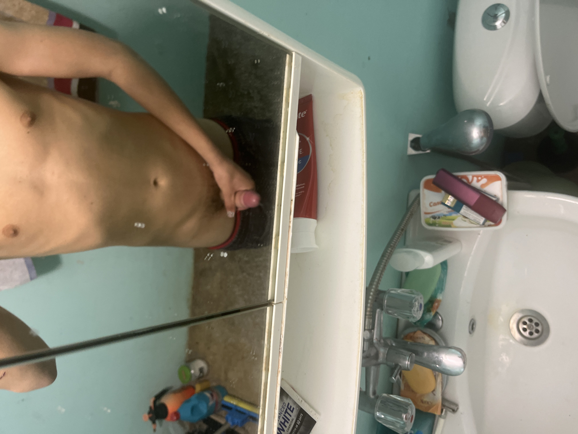 A nude male is reflected in a bathroom mirror. The bathroom has a toilet, sink, and vanity with toiletries. The location is likely Sofia, Bulgaria based on the visual style and common items.