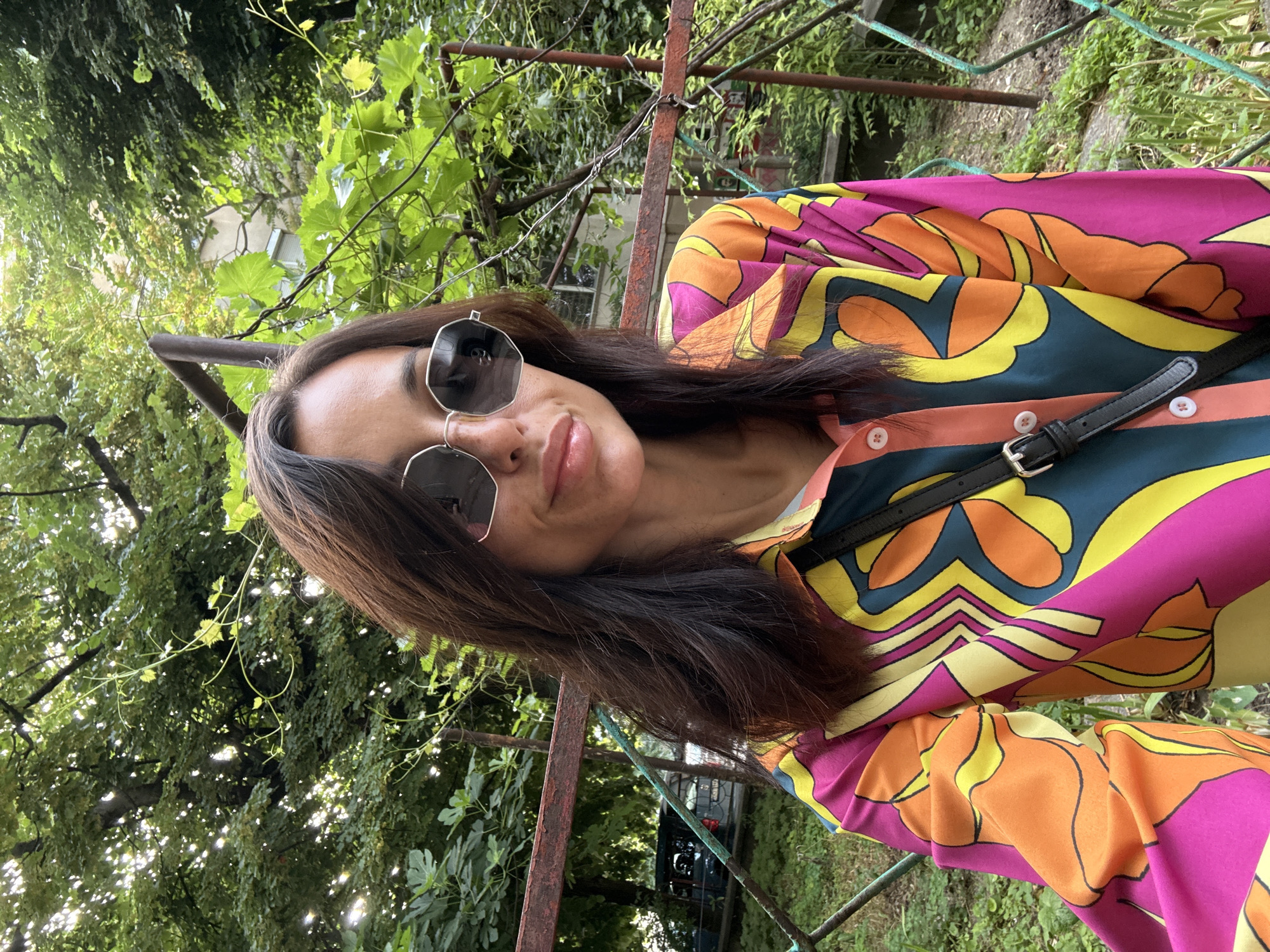 A medium-shot, front-facing selfie of a woman is captured outdoors. She has long, dark brown hair and is wearing large, hexagonal sunglasses. Her lips are slightly parted, and she has a faint smile. She is wearing a vibrantly colored, collared shirt with a psychedelic pattern featuring shades of pink, orange, yellow, and teal. A black strap from a bag is visible across her chest.
The background consists of lush green foliage and trees, suggesting a park or garden setting. A rusty metal fence runs across the mid-ground, with a building visible in the distance. The lighting indicates it is daytime. The context provided indicates the location is in the city of Pleven, Bulgaria.