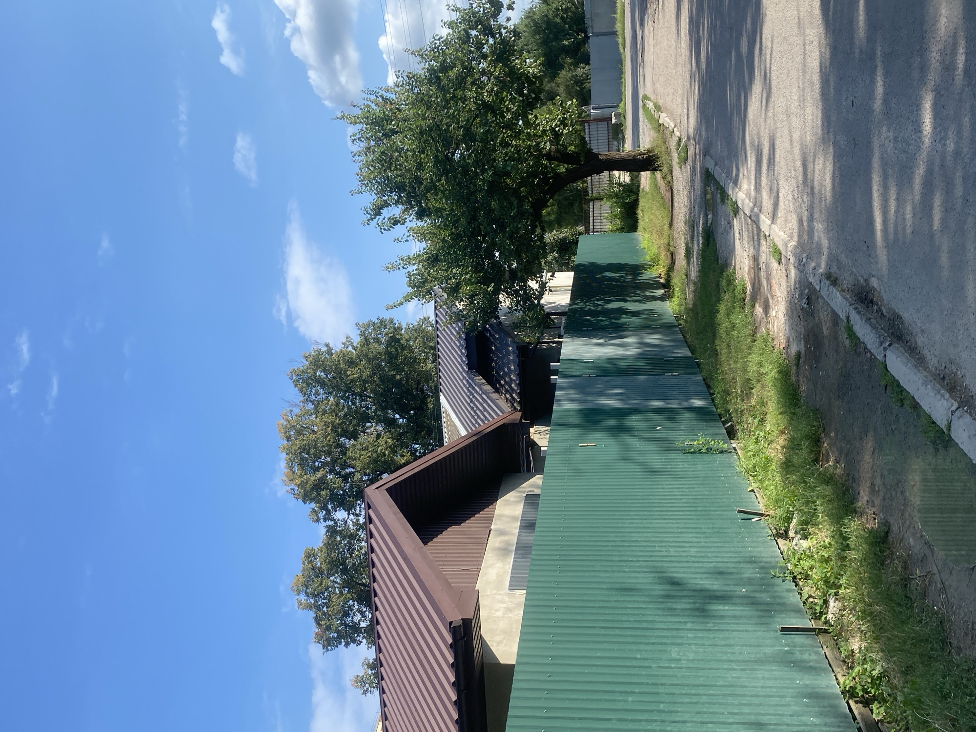 A sunny day with a blue sky and scattered clouds is depicted. A green corrugated metal fence runs along a paved road. To the left of the fence, a section of a building with a brown metal roof is visible. A tree with green leaves stands on the right side of the road, near the fence. The scene is likely in Poltava, Ukraine, given the context.