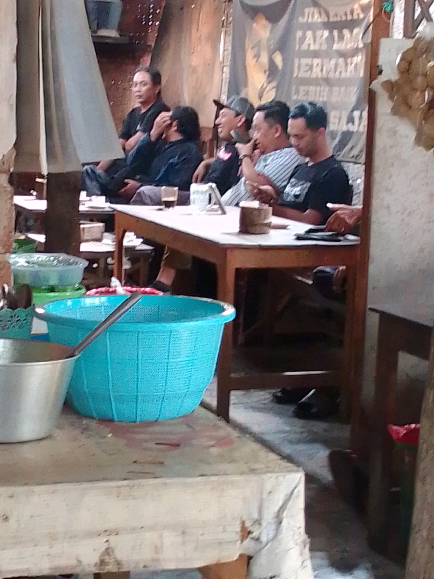 The image depicts a group of men seated around a wooden table, likely at a roadside eatery or warung in Trenggalek, Indonesia. The environment appears casual and somewhat rustic, with a banner displaying text in Indonesian hanging in the background. The men are engaged in conversation or observing something, with one man looking towards the camera. A blue plastic basin, a metal bowl, and various other items are visible on the table and surrounding surfaces, suggesting a meal or drink service. The lighting is dim, possibly indicating an overcast day or an indoor setting with limited natural light.
