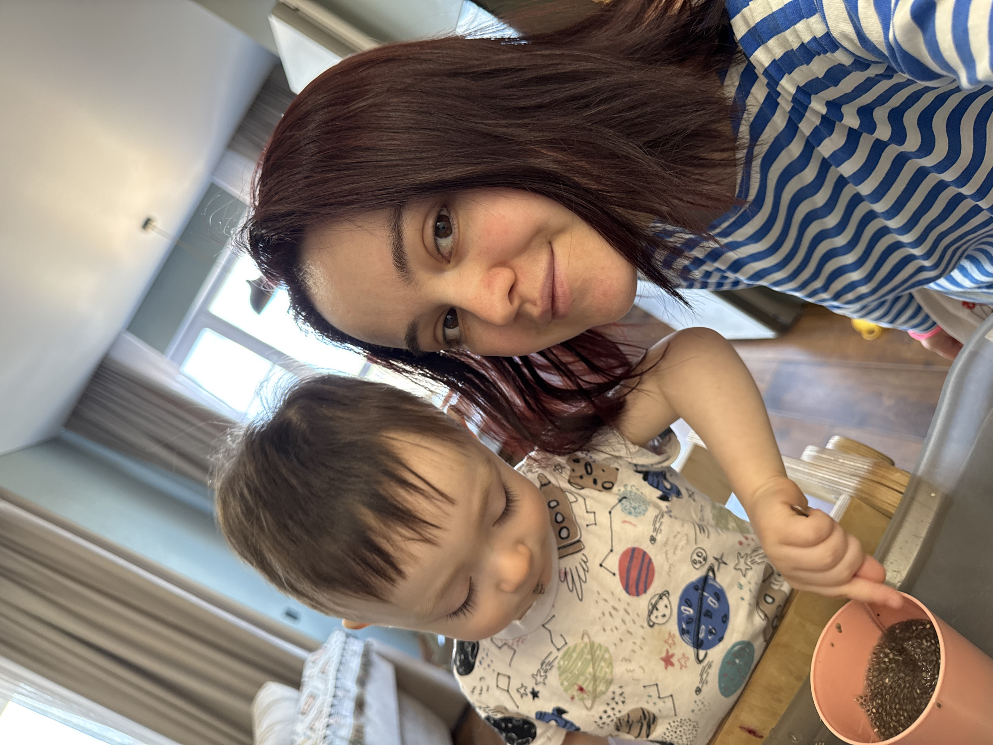 A mother and her young child are captured in a close-up, diagonal photograph taken indoors in Izhevsk, Russia. The woman, likely the mother, is in the upper right portion of the frame, smiling warmly and looking directly at the camera. She has long, dark reddish-brown hair and is wearing a blue and white striped long-sleeved top. Her skin shows some freckles.
Below her, towards the bottom left, is a young child, likely a toddler, deeply engrossed in an activity. The child has light brown hair and is wearing a white t-shirt patterned with colorful space-themed illustrations, including planets, rockets, and stars. The child's gaze is directed downwards at a pink cylindrical container, which holds a quantity of what appears to be chia seeds mixed with liquid. The child is holding a small, metallic utensil, possibly a spoon, and appears to be stirring or exploring the contents of the cup.
The setting is an indoor environment, likely a home, with natural light streaming in from a large window visible in the background, partially covered by light-colored curtains. A white wall and a ceiling are also visible. The activity takes place on a light-colored wooden surface, possibly a table or high chair tray. The overall mood is intimate and suggests a moment of shared activity or play between the mother and child. There is no visible text in the image.