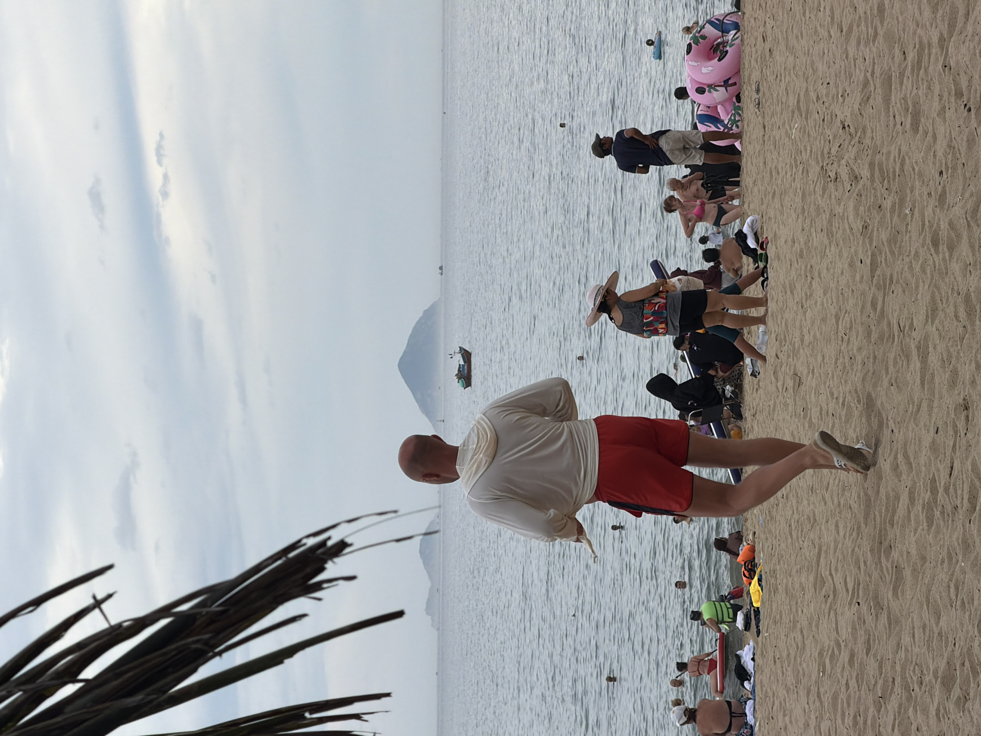 The image shows a cloudy day at Nha Trang beach in Vietnam. In the foreground, a bald man wearing a white hooded sweatshirt and red swim trunks is walking away from the camera. The beach is populated with people swimming and sunbathing. Several inflatable pink rings are stacked near the right side of the frame. In the distance, boats are visible on the ocean, and islands can be seen on the horizon. A palm frond hangs over the top left corner of the image.